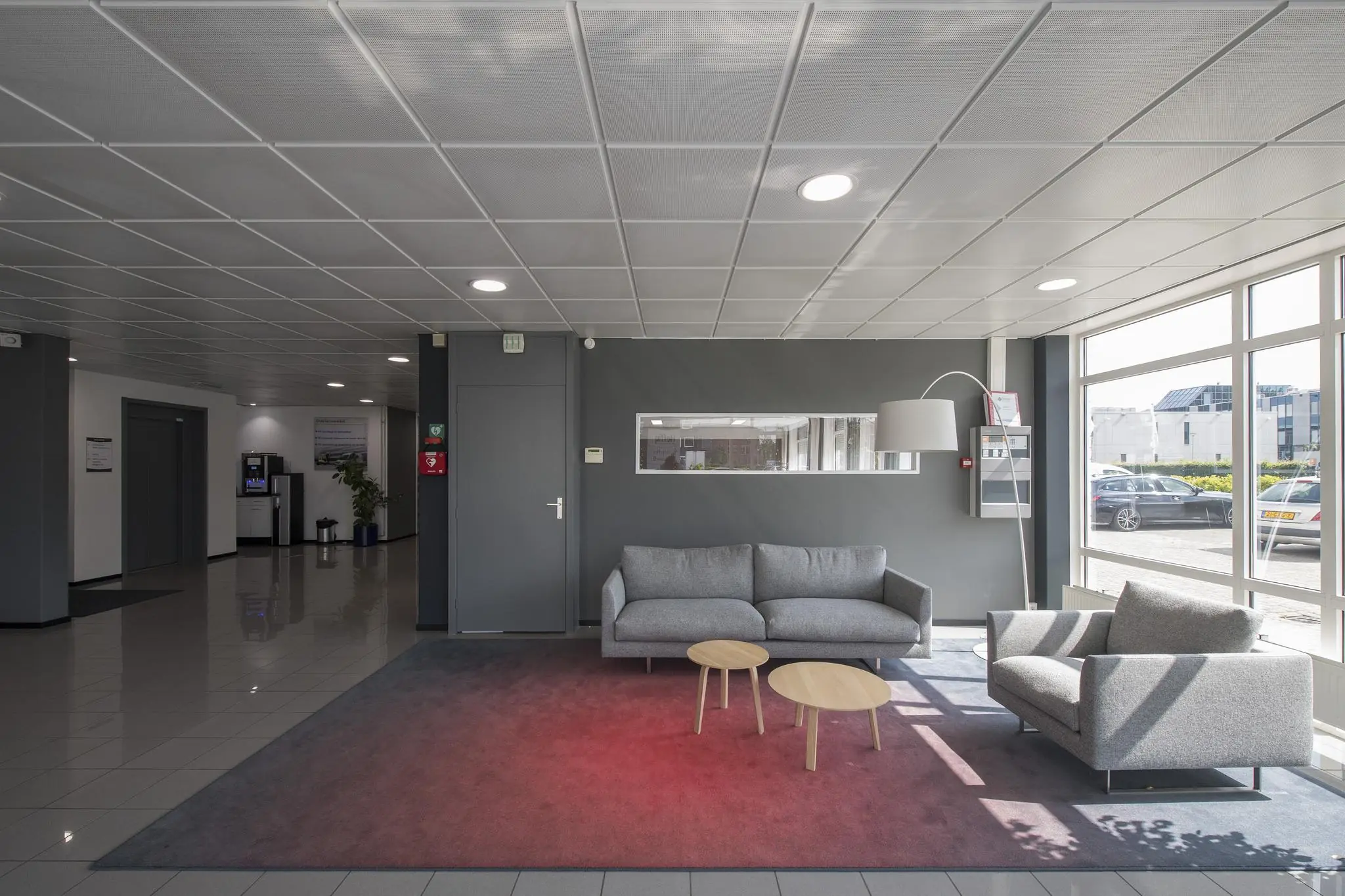 Moderne wachtruimte met grijze zitbanken, houten tafeltjes en grote ramen die uitzicht bieden op een bedrijventerrein.