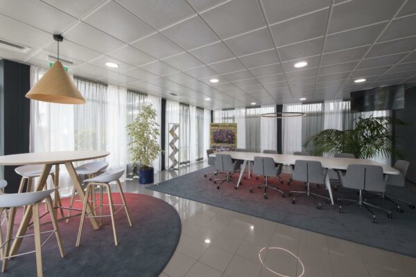 Moderne vergaderruimte met een lange tafel, grijze stoelen op wieltjes, hoge krukken aan een ronde tafel en grote ramen met witte gordijnen.