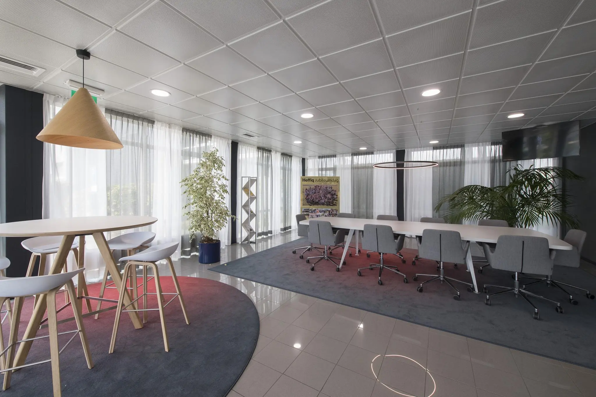 Moderne vergaderruimte met een lange tafel, grijze stoelen op wieltjes, hoge krukken aan een ronde tafel en grote ramen met witte gordijnen.