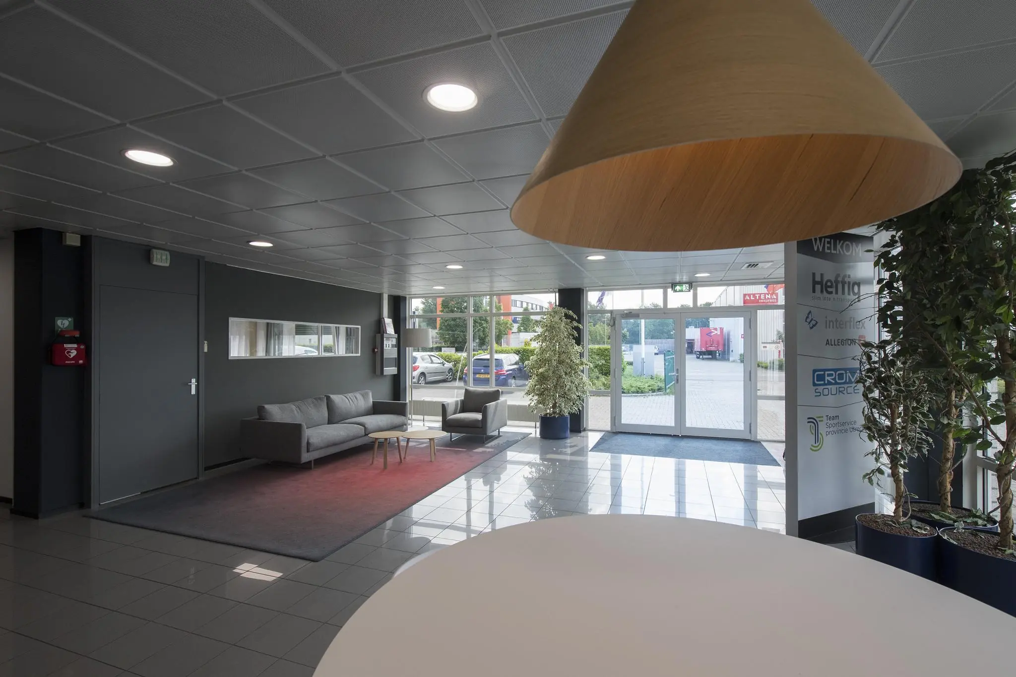Moderne bedrijfsentree met zithoek, grote hanglamp, planten en glazen deuren naar buiten bij Witboom.