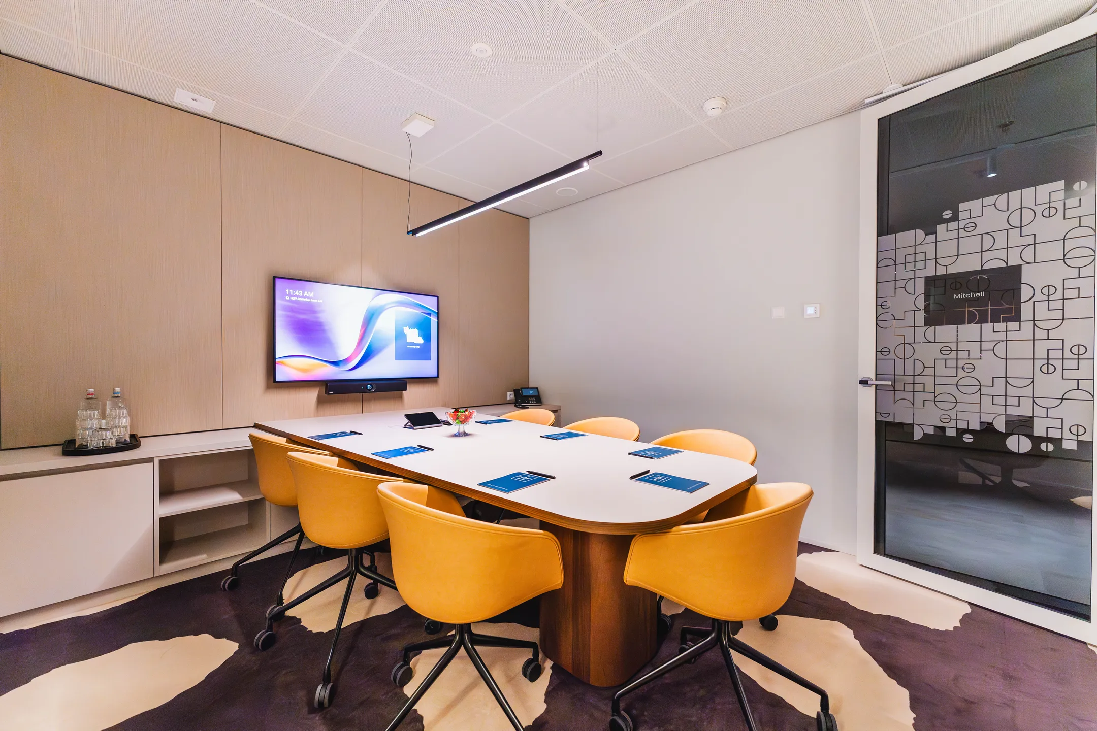 Een moderne vergaderruimte met een witte tafel, oranje stoelen, een scherm aan de muur en de naam "Mitchell" op de deur.
