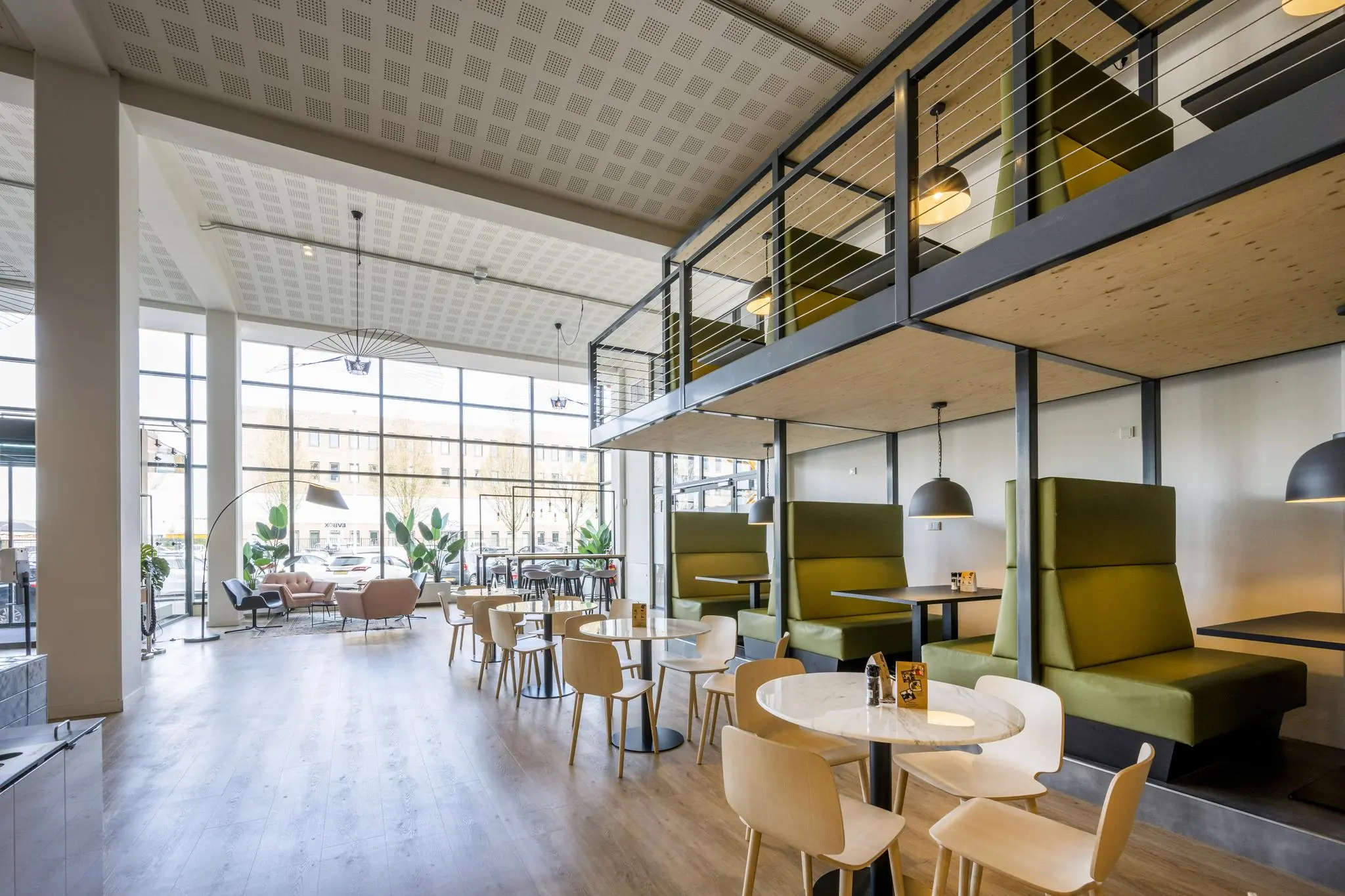 Modern ingerichte horecaruimte aan de Kabelweg met hoge ramen, houten vloeren en groene zitbanken in een mezzanine-opstelling.