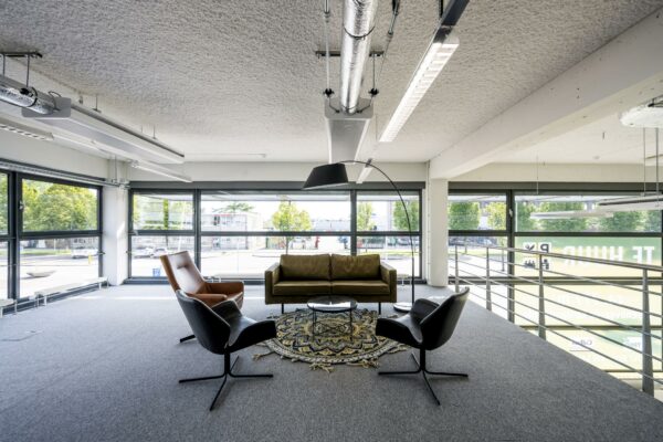 Moderne zitruimte op een kantoor aan de Kabelweg met een bank, stoelen, kleed en grote ramen met uitzicht op de straat.
