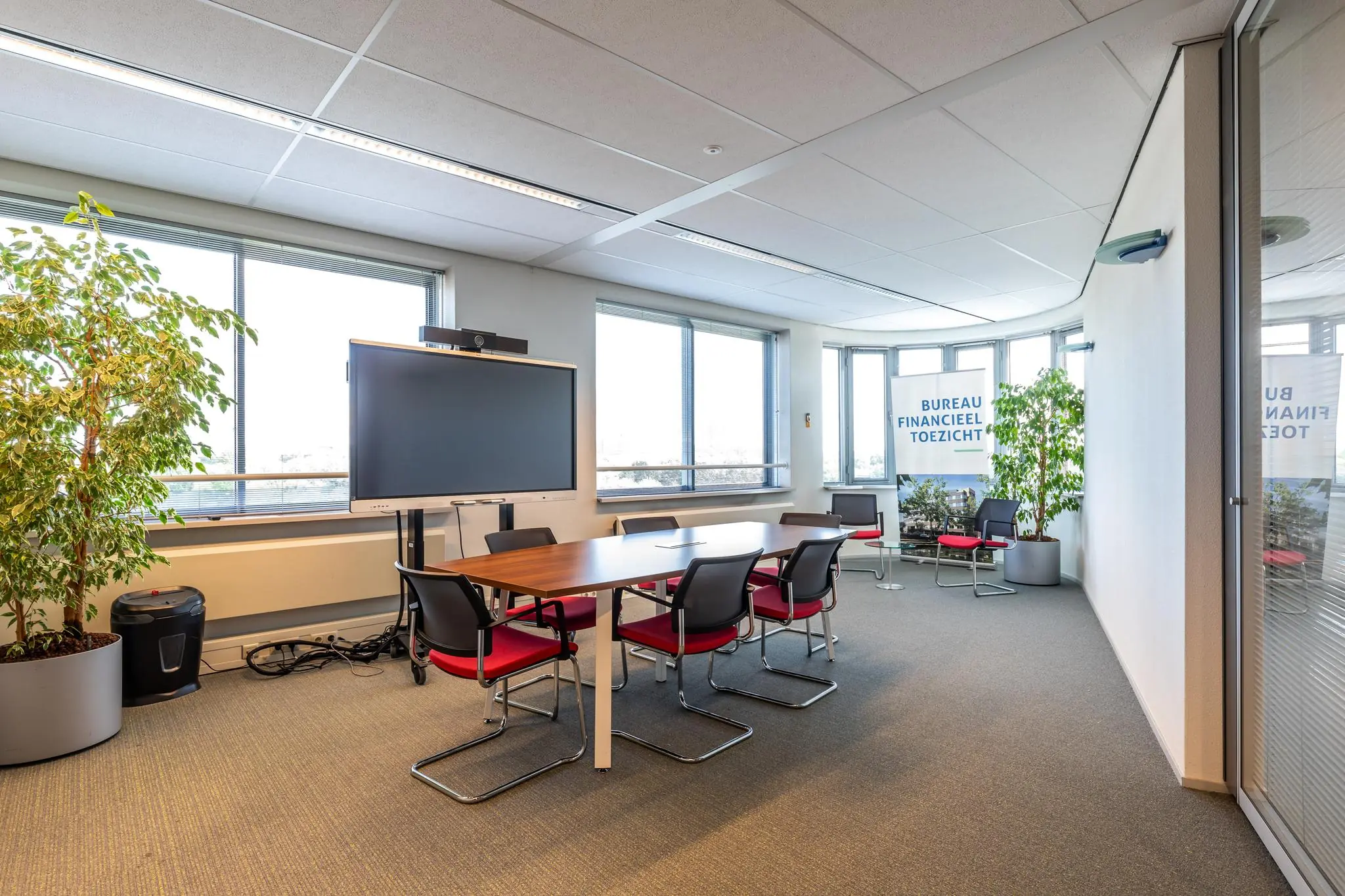 Vergaderruimte met een groot scherm, zes stoelen rond een tafel en zicht op het Bureau Financieel Toezicht.