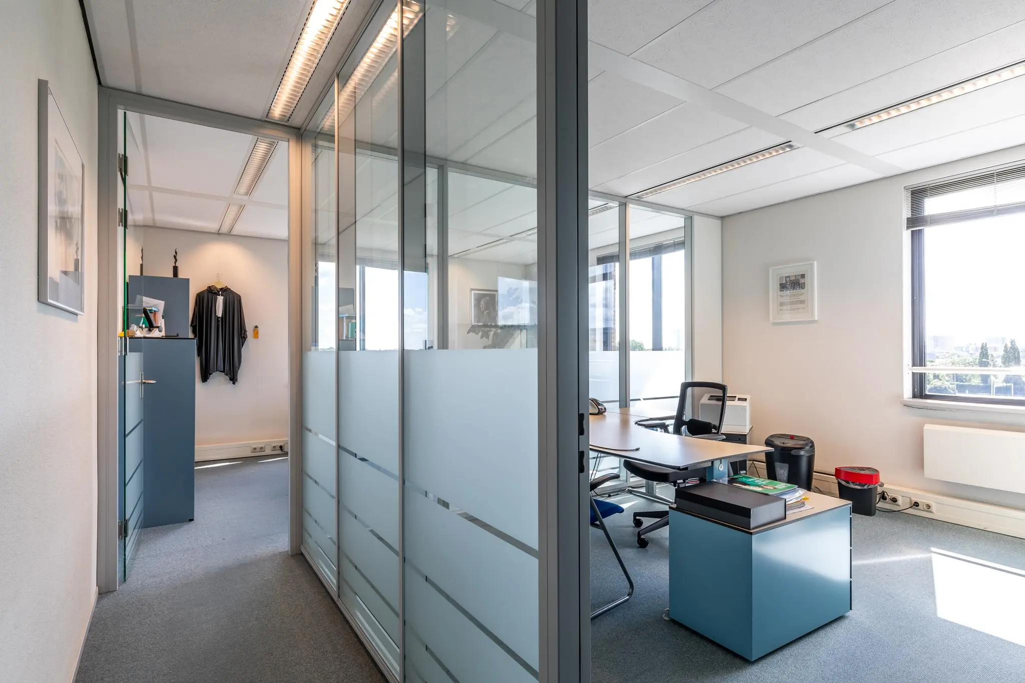 Modern kantoor met glazen wanden aan de Euclideslaan, ingericht met een bureau, stoelen en een toga aan de muur.