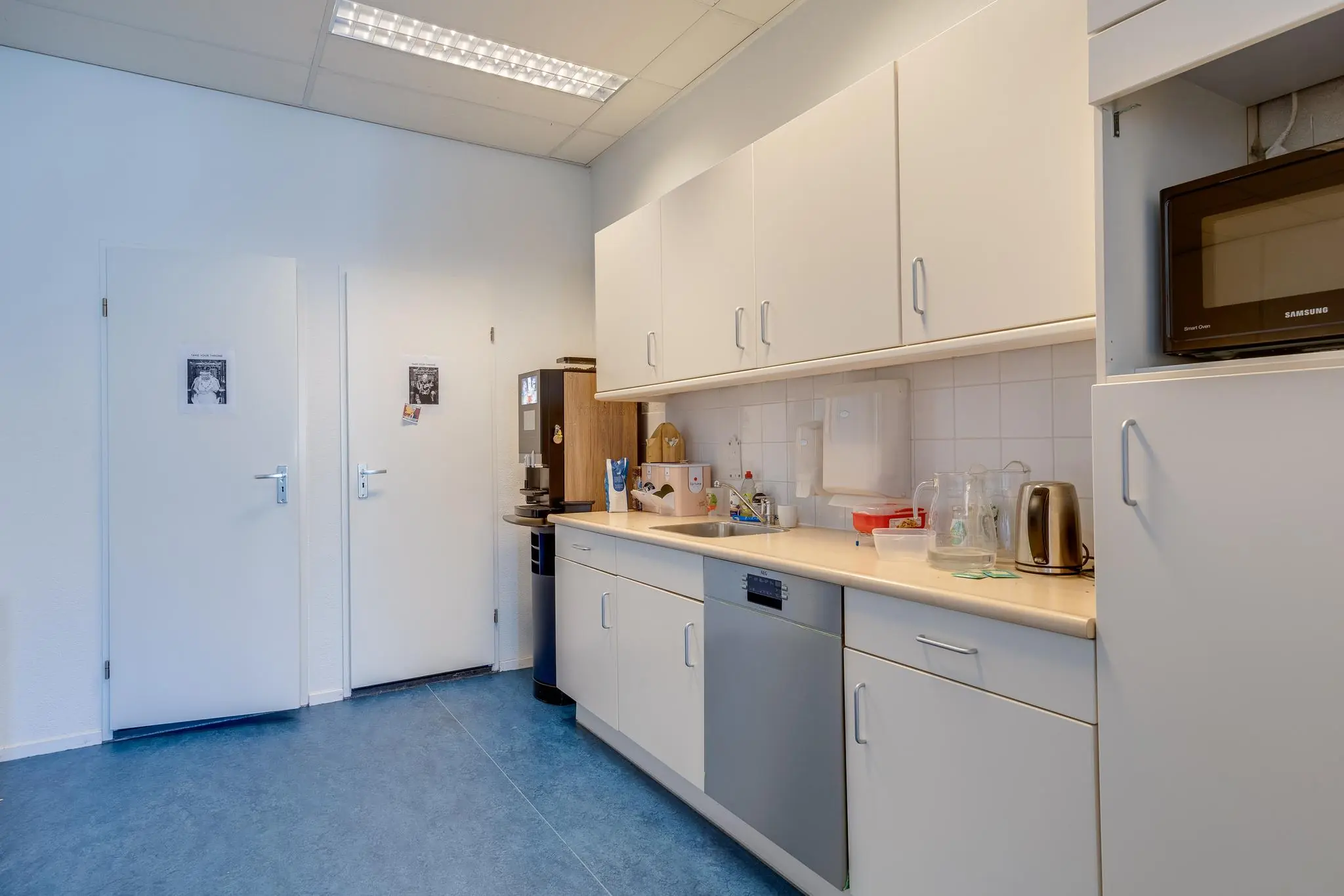 Kantoorkeuken met witte kastjes, een aanrecht met vaatwasser, waterkoker, koffiezetapparaat en magnetron.