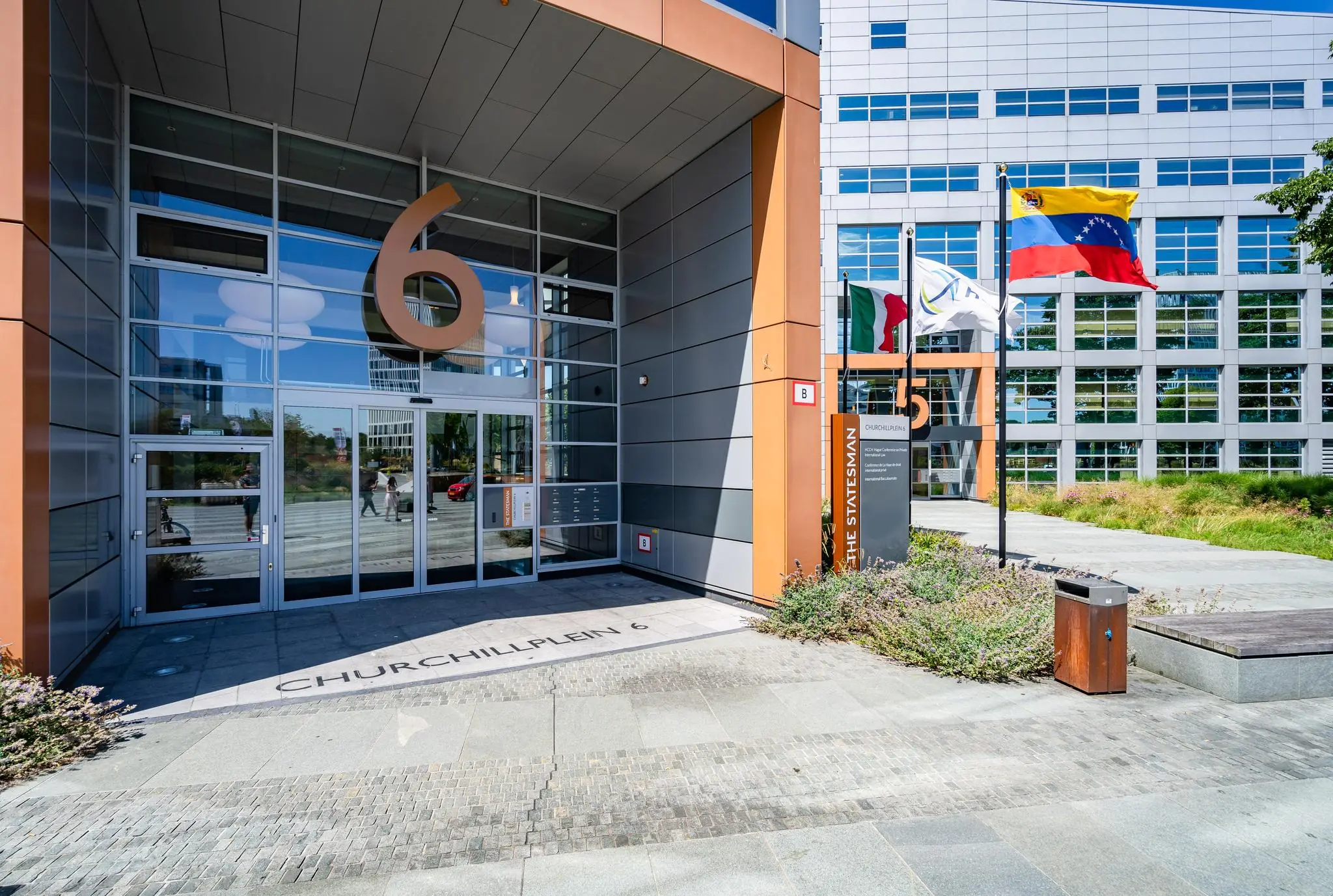 Ingang van kantoorgebouw Churchillplein 6 in Den Haag met grote huisnummers, vlaggen en het bord "The Statesman".