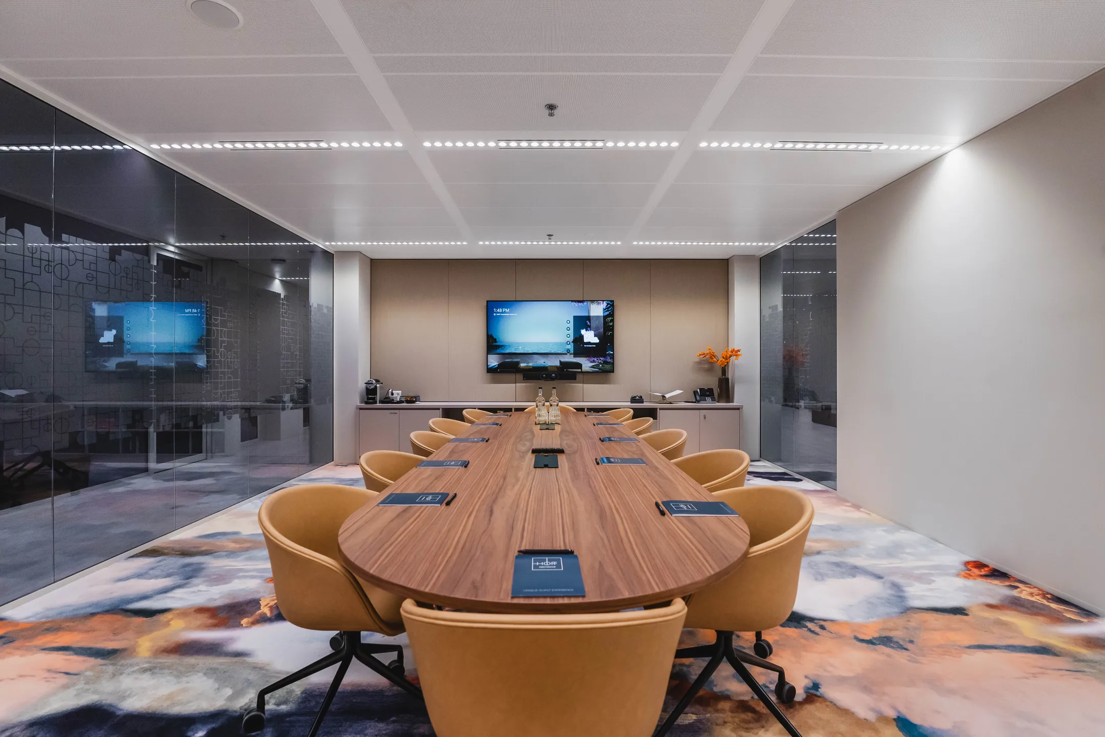 Een moderne vergaderruimte met een lange houten tafel, omringd door beige stoelen en voorzien van een scherm aan de muur.