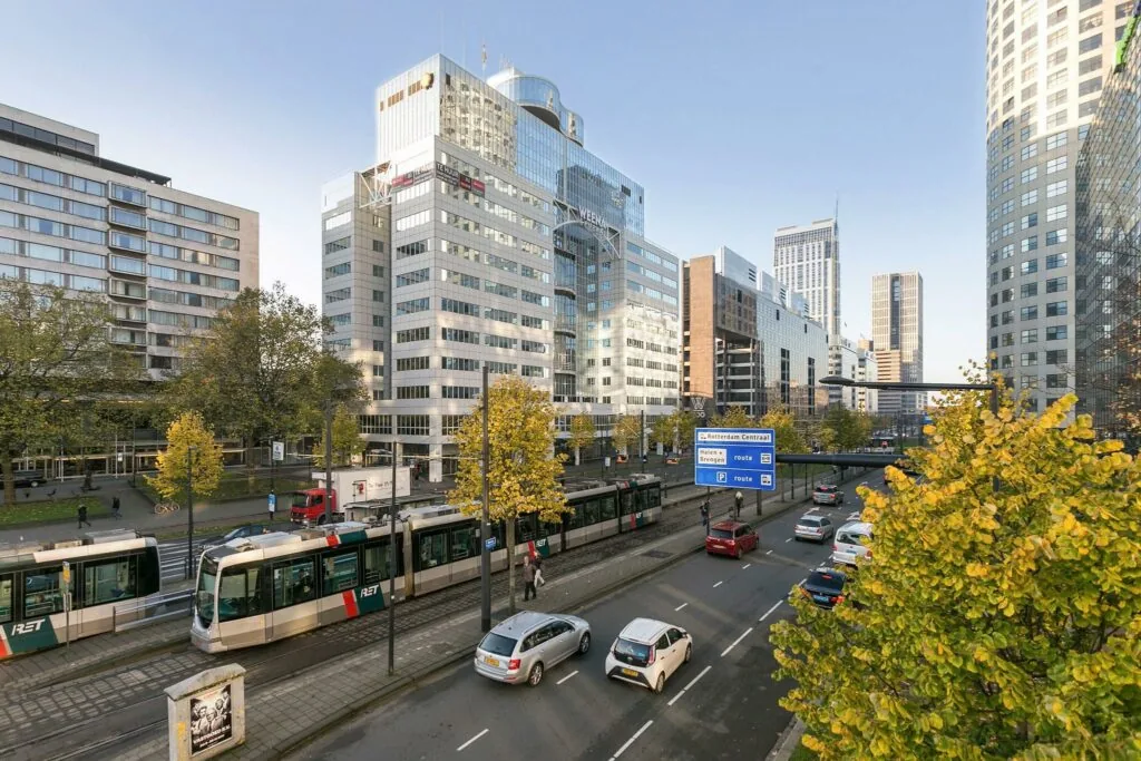 Drukke stadsstraat Weena in Rotterdam met trams, auto's, moderne kantoorgebouwen en bewegwijzering richting Rotterdam Centraal.