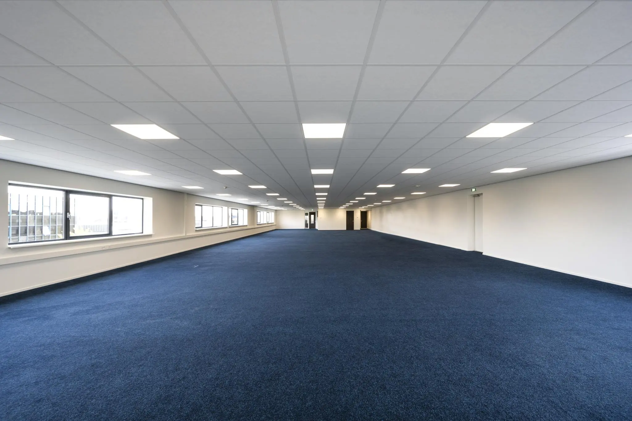 Ruime, lege kantoorruimte op de Dynamostraat met blauw tapijt, witte plafonds en grote ramen langs de zijmuur.