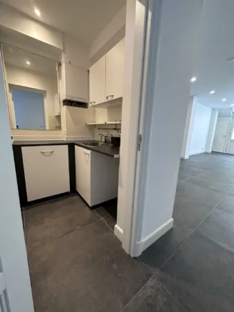 Moderne, compacte bijkeuken met witte kasten en een zwart werkblad, grenzend aan een ruime kamer met donkergrijze vloertegels.