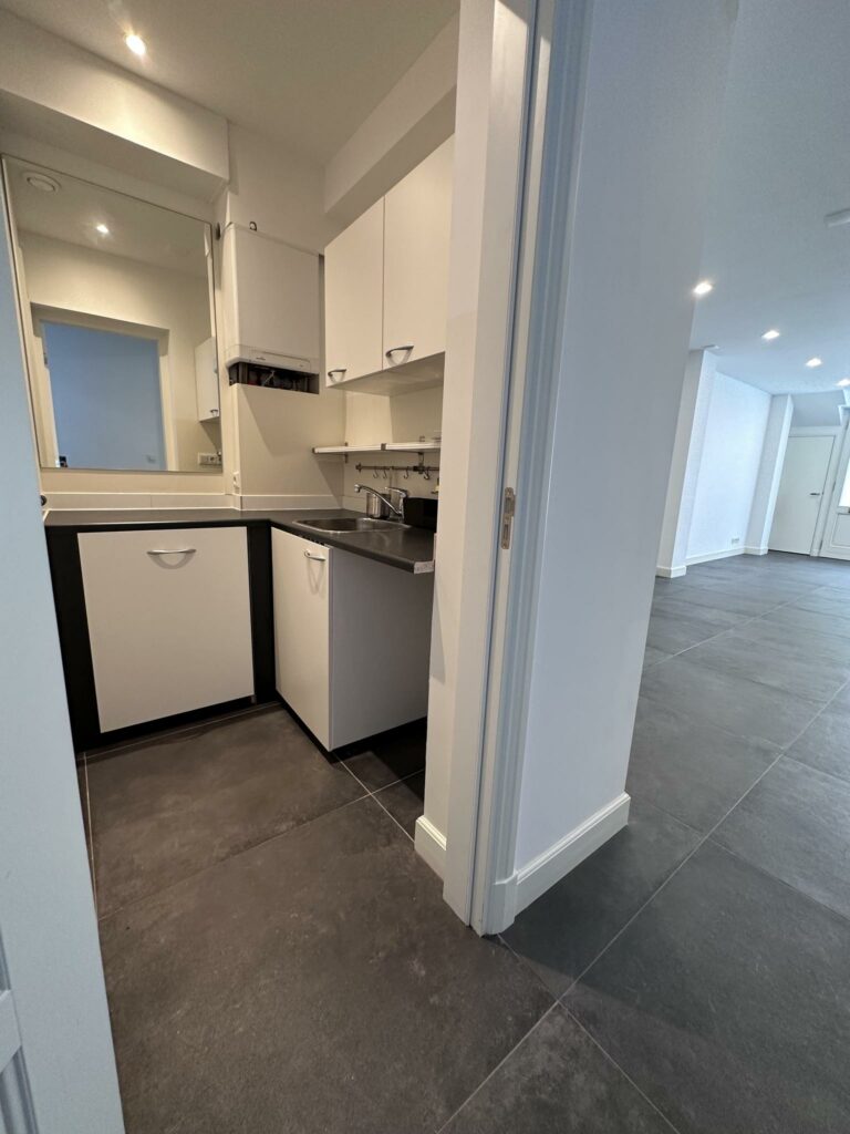 Moderne, compacte bijkeuken met witte kasten en een zwart werkblad, grenzend aan een ruime kamer met donkergrijze vloertegels.