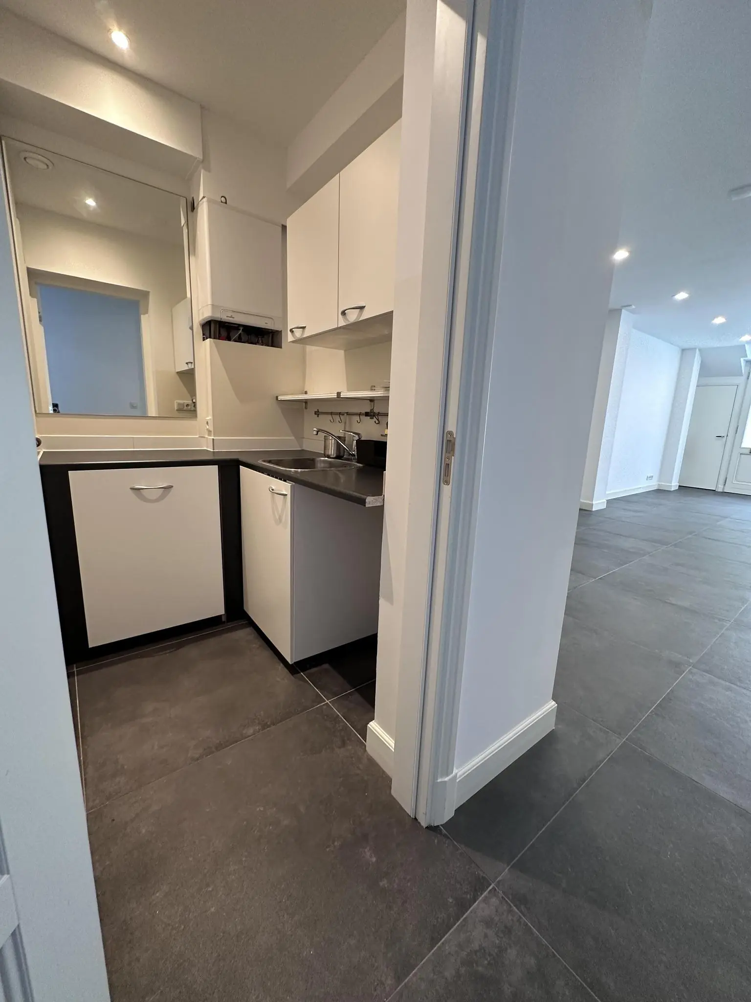 Moderne, compacte bijkeuken met witte kasten en een zwart werkblad, grenzend aan een ruime kamer met donkergrijze vloertegels.