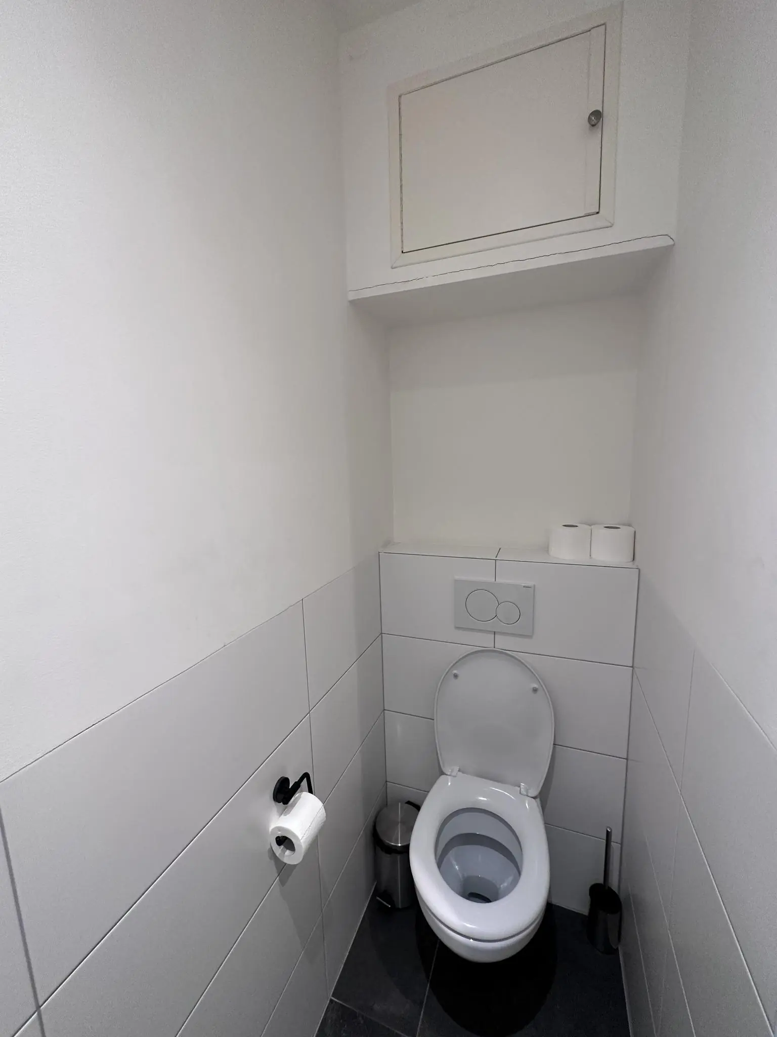 Kleine, witte toiletruimte met hangtoilet, toiletrolhouder, prullenbak, toiletborstel en opbergkastje bovenin.