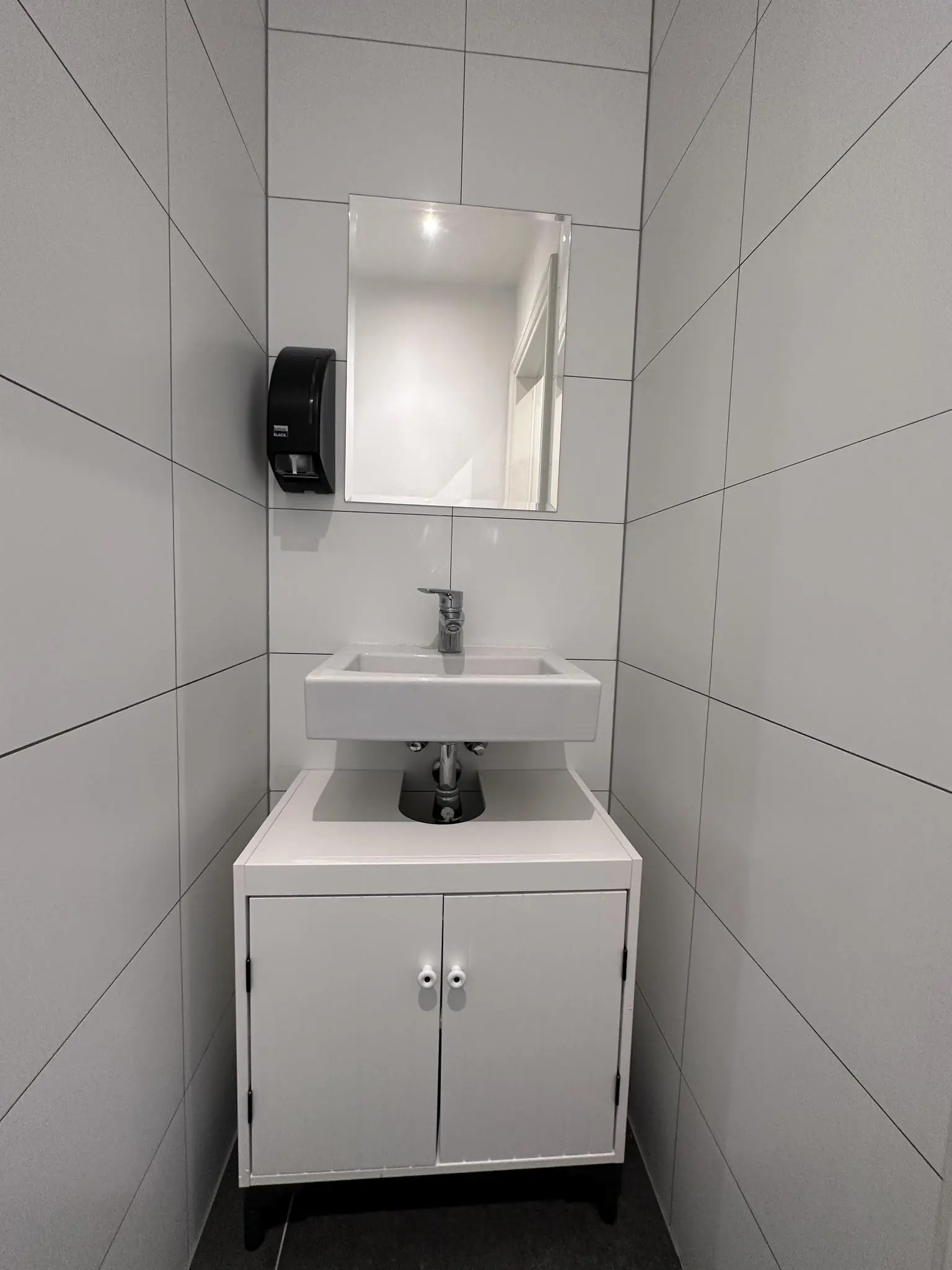 Wit betegelde toiletruimte met wastafel, spiegel, onderkast en zwarte muurdispenser.