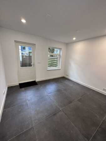 Leeg, modern afgewerkte kamer met grote donkere vloertegels, witte muren en een deur met raam aan de Ceintuurbaan.