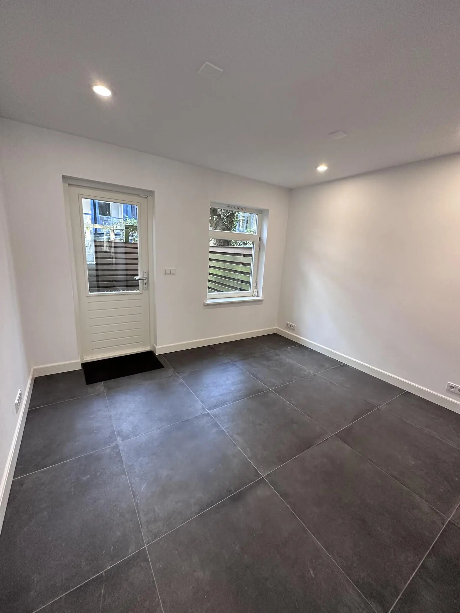 Leeg, modern afgewerkte kamer met grote donkere vloertegels, witte muren en een deur met raam aan de Ceintuurbaan.