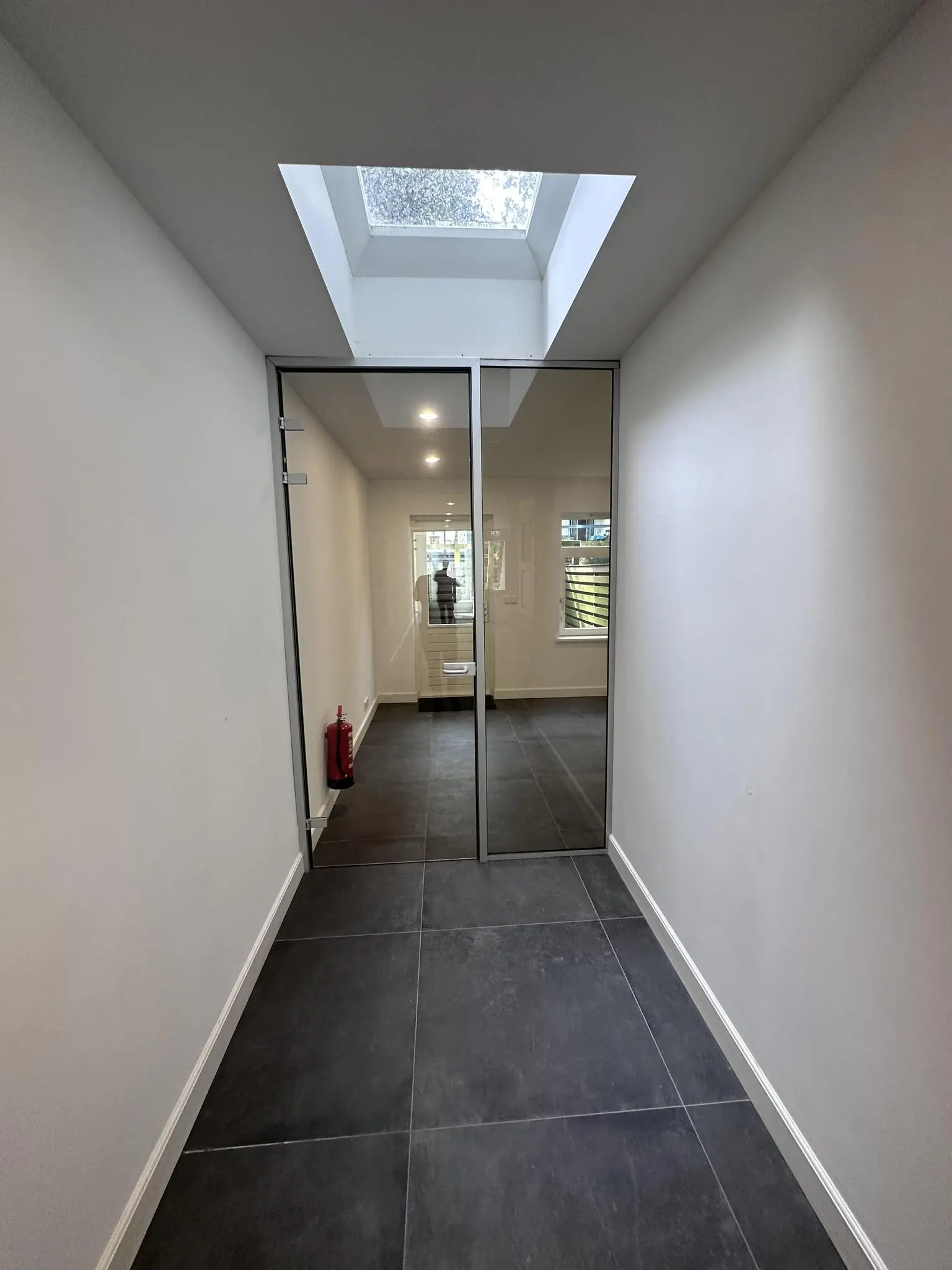 Moderne gang met donkergrijze vloertegels, een glazen deur, dakraam en een brandblusser in een pand aan de Ceintuurbaan.