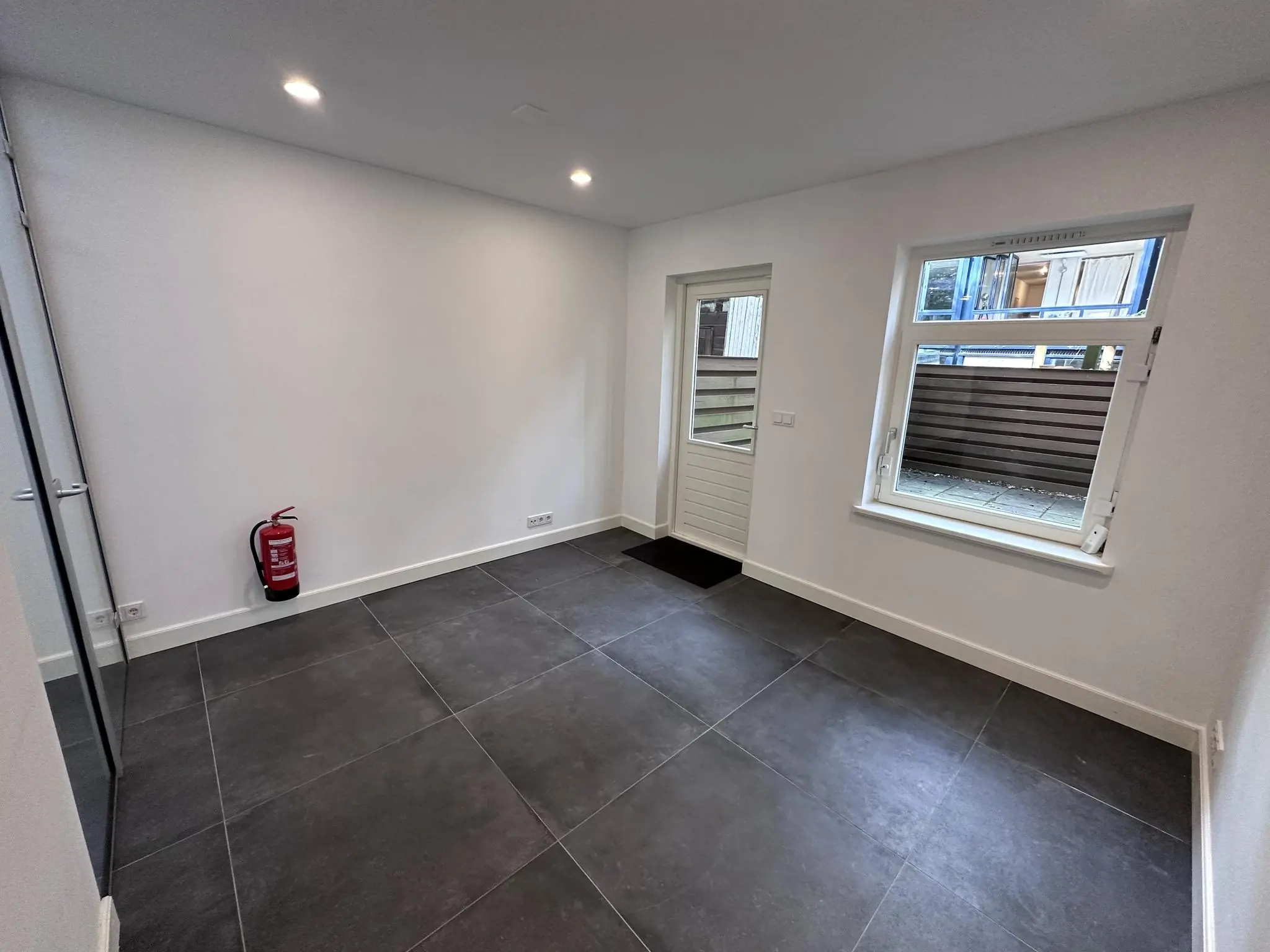 Lege kamer met donkere vloertegels, een witte muur, raam, deur naar buiten en een rode brandblusser in de hoek.