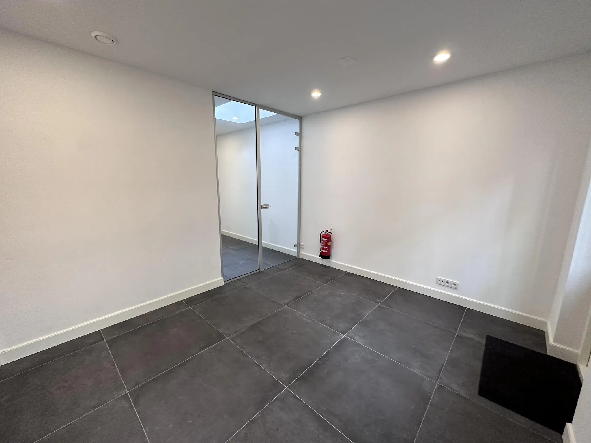 Kleine lege kamer met een glazen deur, zwarte vloertegels en een brandblusser tegen de witte muur.