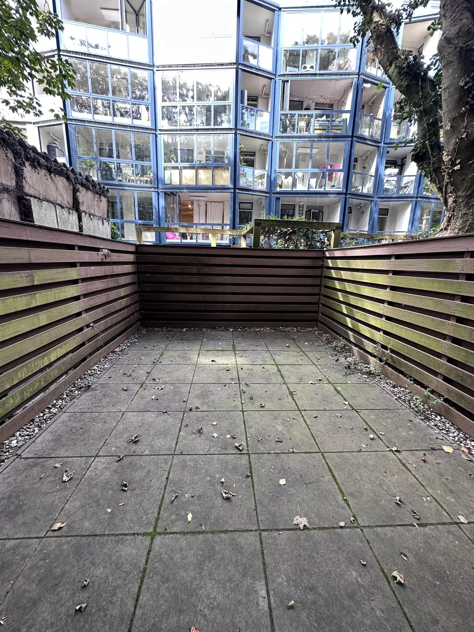 Afgesloten betegeld terras met houten schutting en uitzicht op glazen gevel van appartementencomplex aan de Ceintuurbaan.