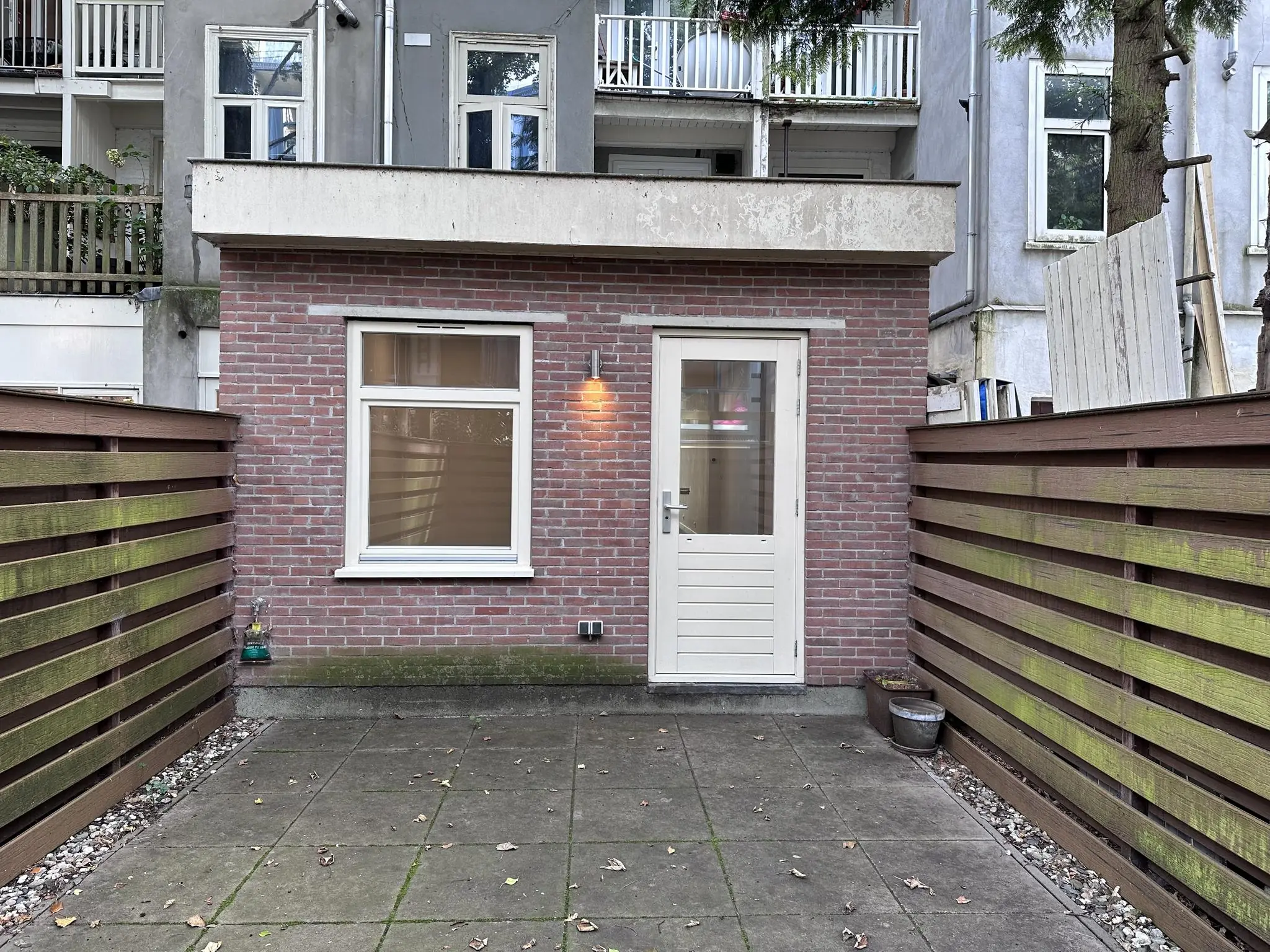 Kleine rode bakstenen uitbouw met witte deur en raam, gelegen aan de Ceintuurbaan met houten schuttingen aan beide zijden.