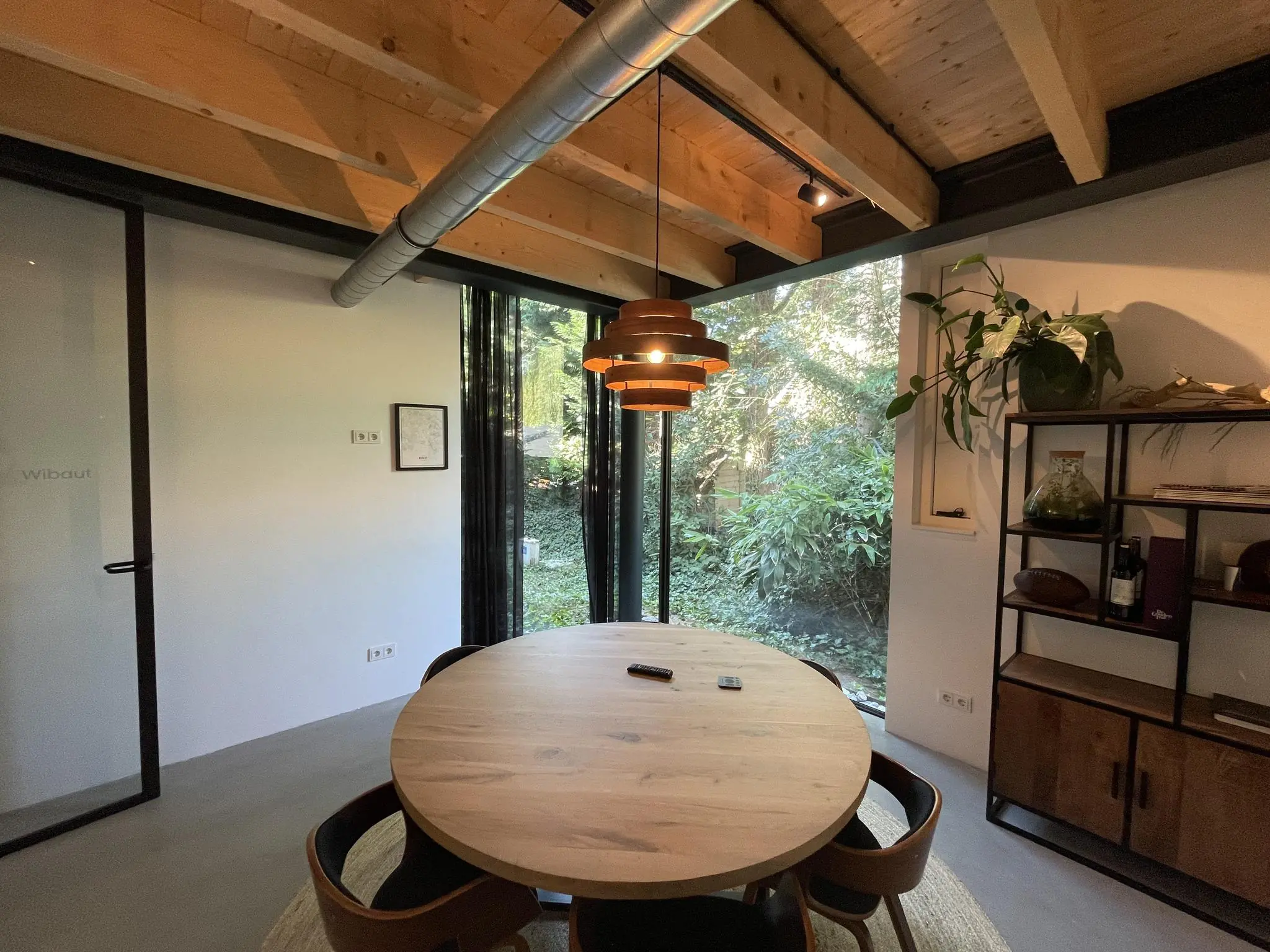 Moderne vergaderruimte aan de Sloterweg met ronde houten tafel, hanglamp, glazen wand en zicht op groene tuin.