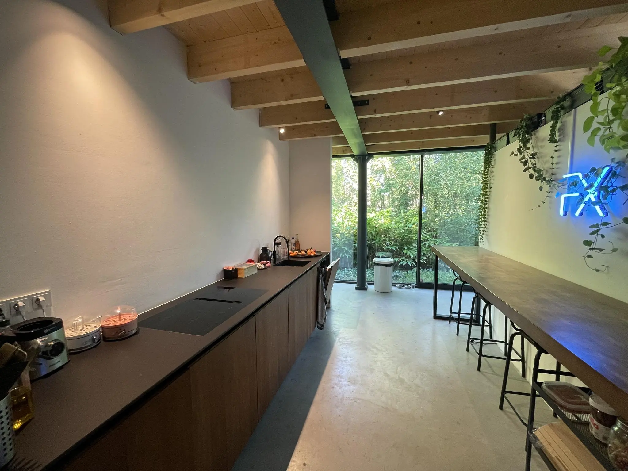 Moderne keuken met houten balkenplafond, lange werktafel, neonverlichting en uitzicht op een groene tuin aan de Sloterweg.