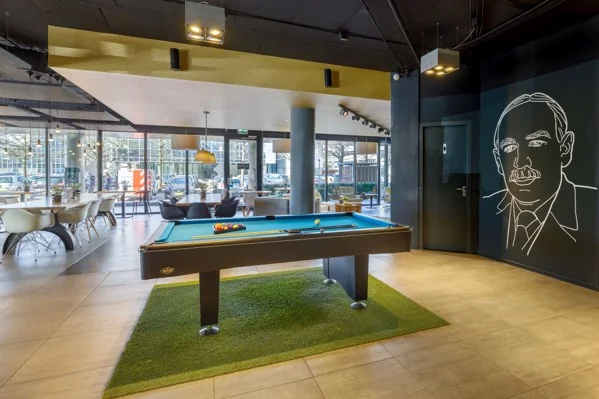 Moderne lounge met een pooltafel op kunstgras, grote ramen en muurtekening van een man bij Hogehilweg.