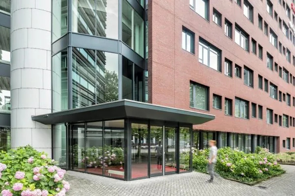 Moderne glazen entree van een kantoorgebouw aan de Hullenbergweg met rode bakstenen gevel en bloeiende hortensia’s.