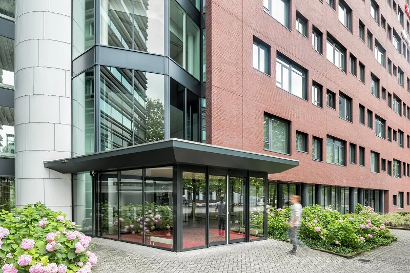 Moderne glazen entree van een kantoorgebouw aan de Hullenbergweg met rode bakstenen gevel en bloeiende hortensia’s.
