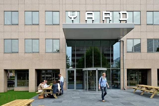 Voor het kantoorgebouw YARD aan de Karspeldreef zitten mensen aan picknicktafels, terwijl iemand het gebouw uitloopt.