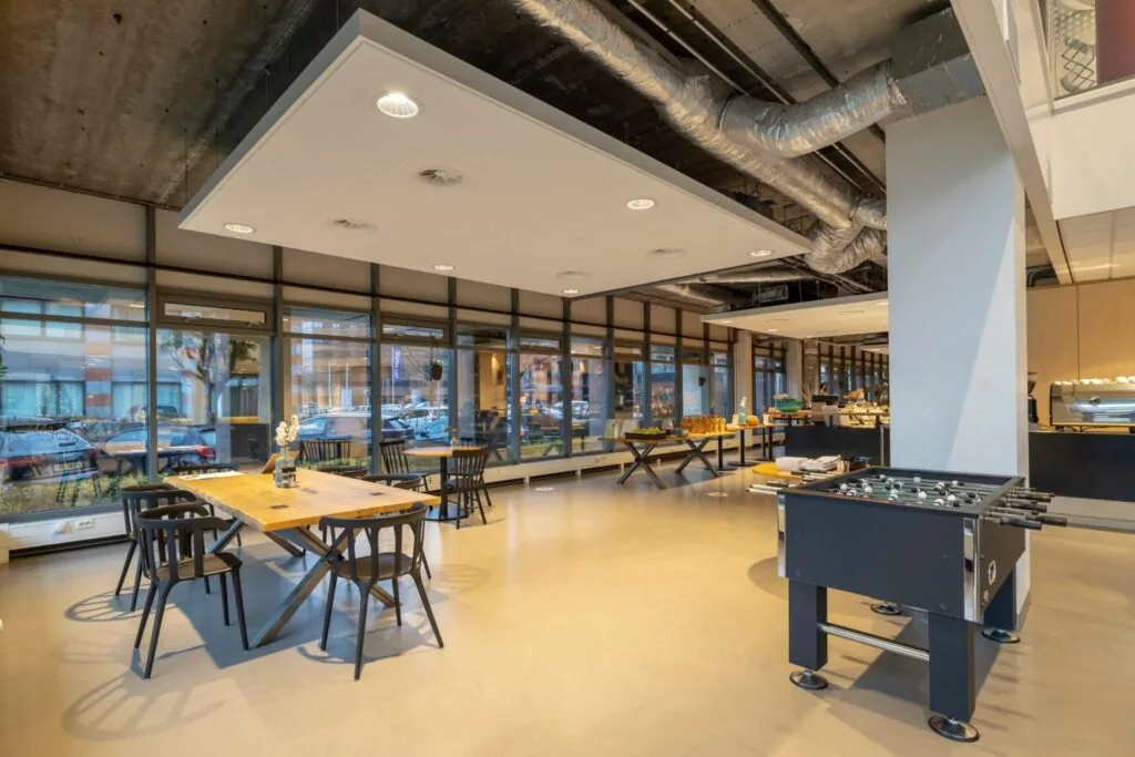 Moderne bedrijfsruimte aan de Karspeldreef met zithoeken, een tafelvoetbaltafel en grote ramen met uitzicht op geparkeerde auto's.