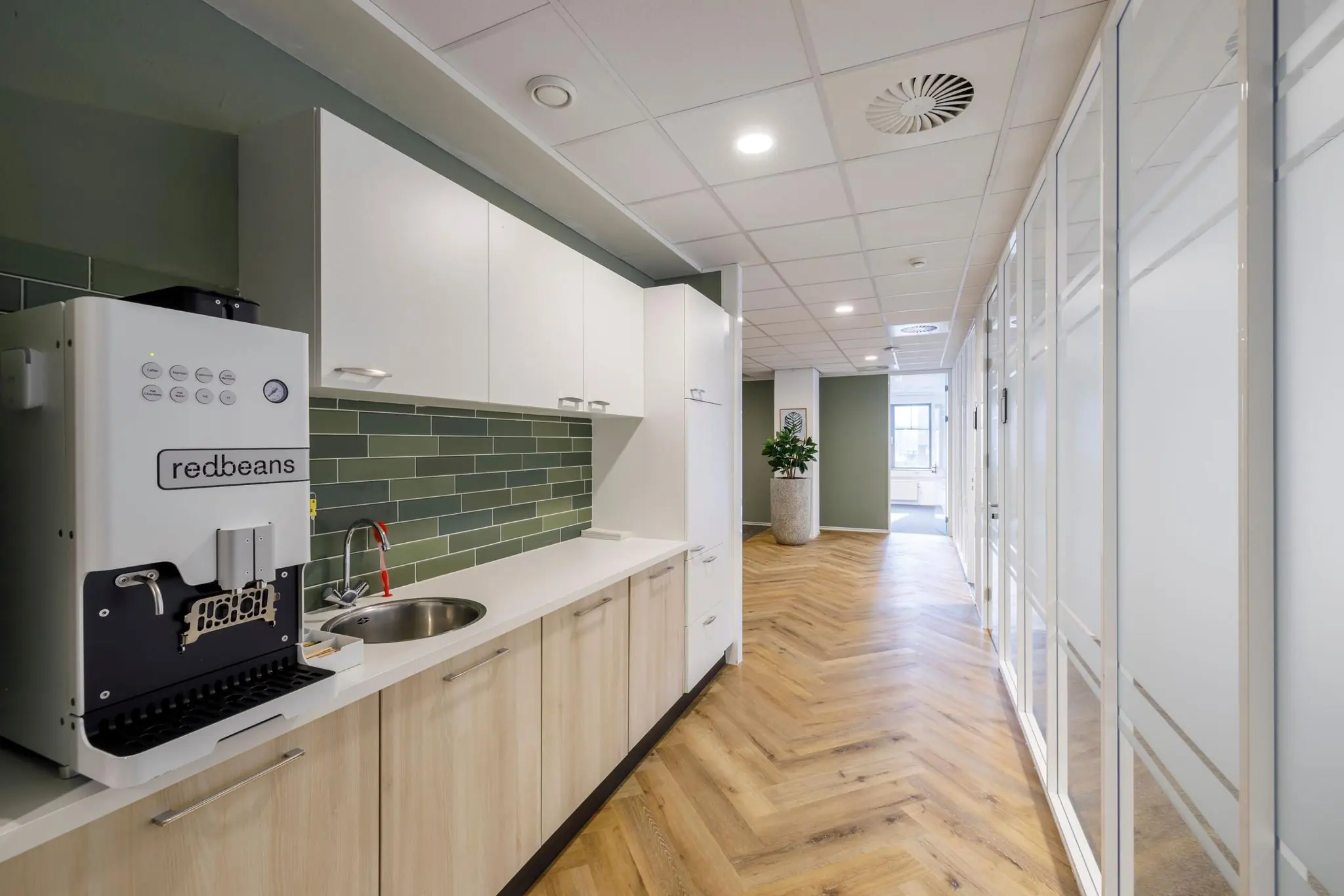 Moderne kantoorkeuken met een Redbeans koffiemachine, groen betegelde achterwand en aangrenzende gang met glaswanden.