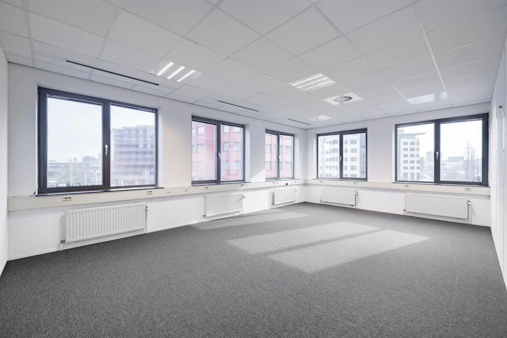 Lichte, lege kantoorruimte aan de Karspeldreef met grote ramen en grijze tapijtvloer.