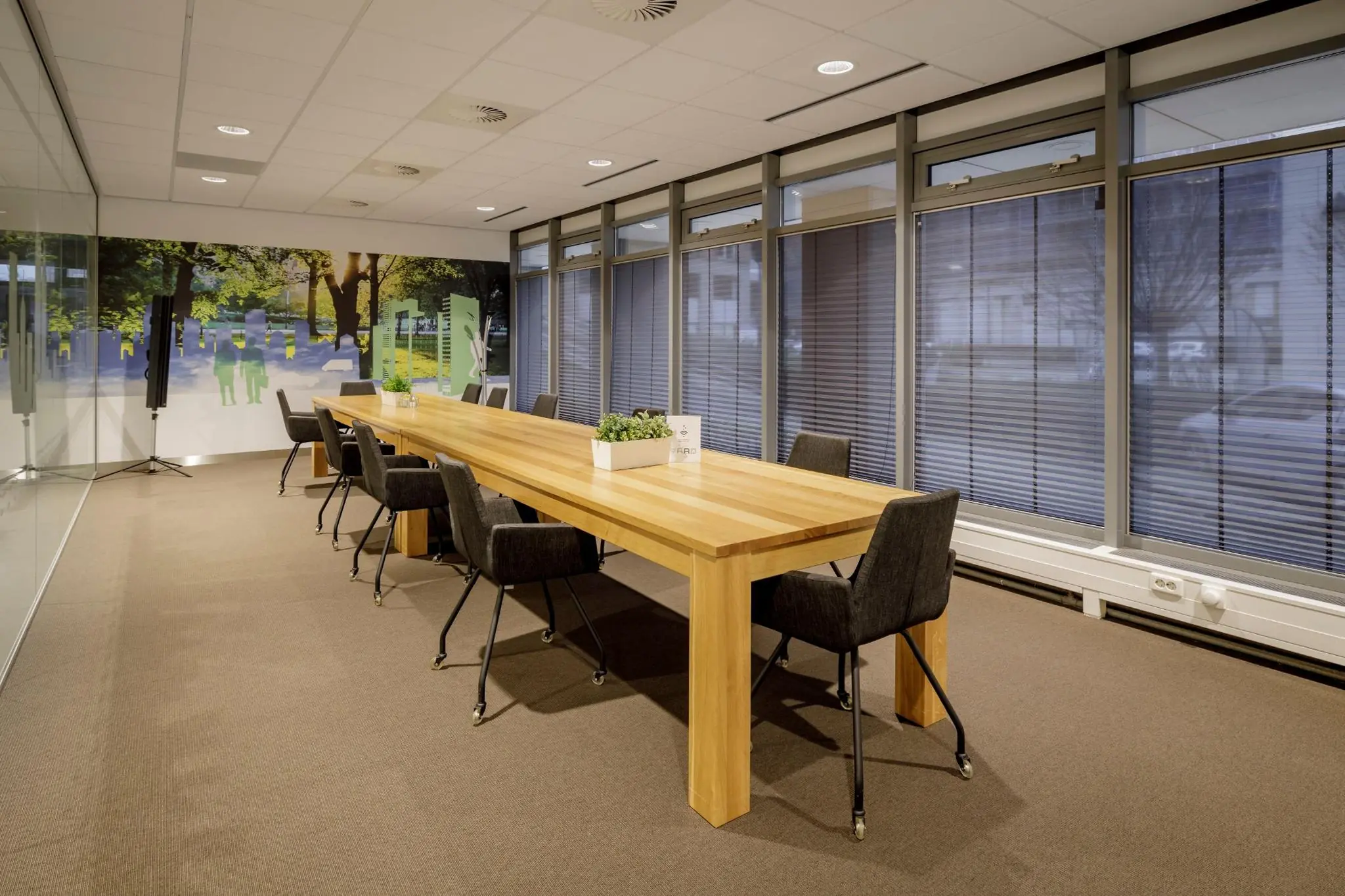 Vergaderruimte aan de Karspeldreef met een lange houten tafel, zwarte stoelen op wieltjes en een kleurrijke muurdecoratie.