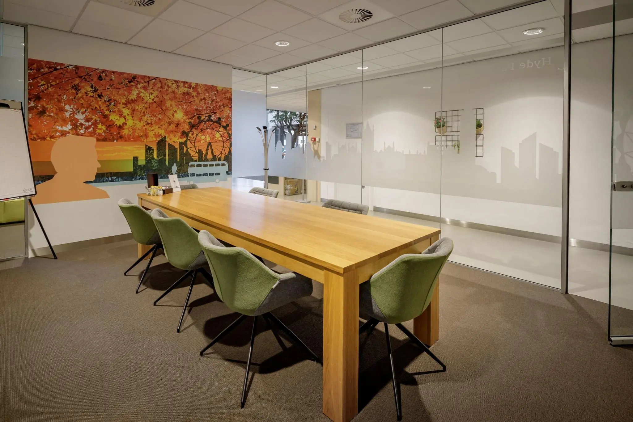 Vergaderruimte aan de Karspeldreef met houten tafel, groene stoelen en een kleurrijke muurschildering met stads- en herfstmotief.