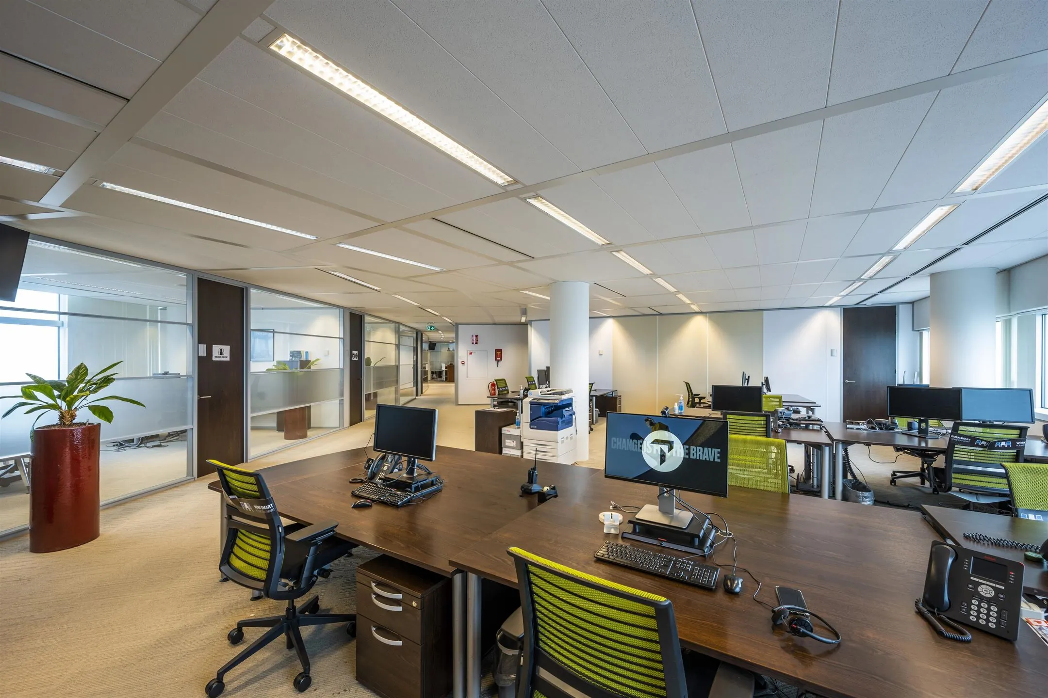 Moderne open kantoorruimte met werkplekken, computers, een printer en groene bureaustoelen.