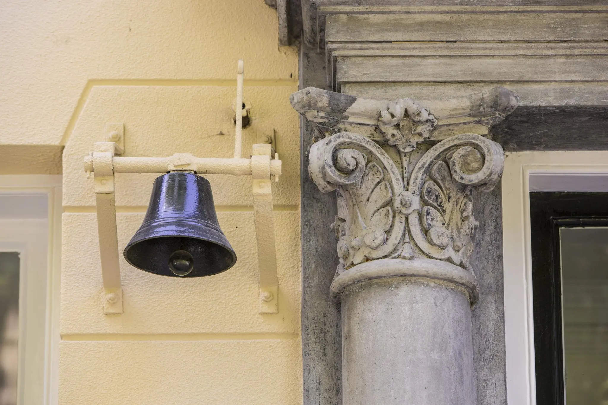 Zwarte metalen bel en sierlijke zuil aan de gevel van een gebouw aan de Naarderstraat.