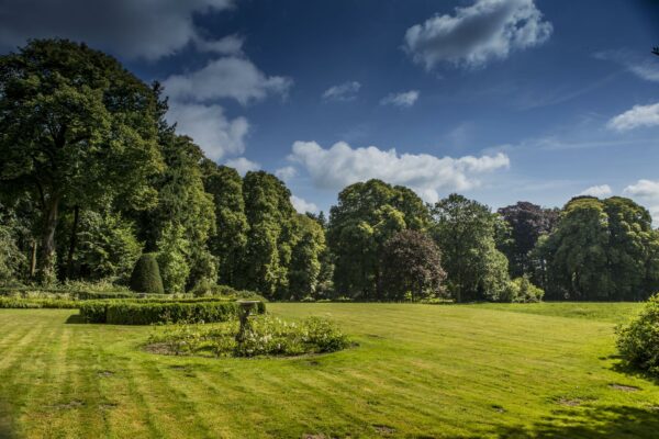 Landschapsfoto van een strak gemaaid gazon met sierlijke heggen en een fontein, omringd door hoge bomen aan de Naarderstraat.