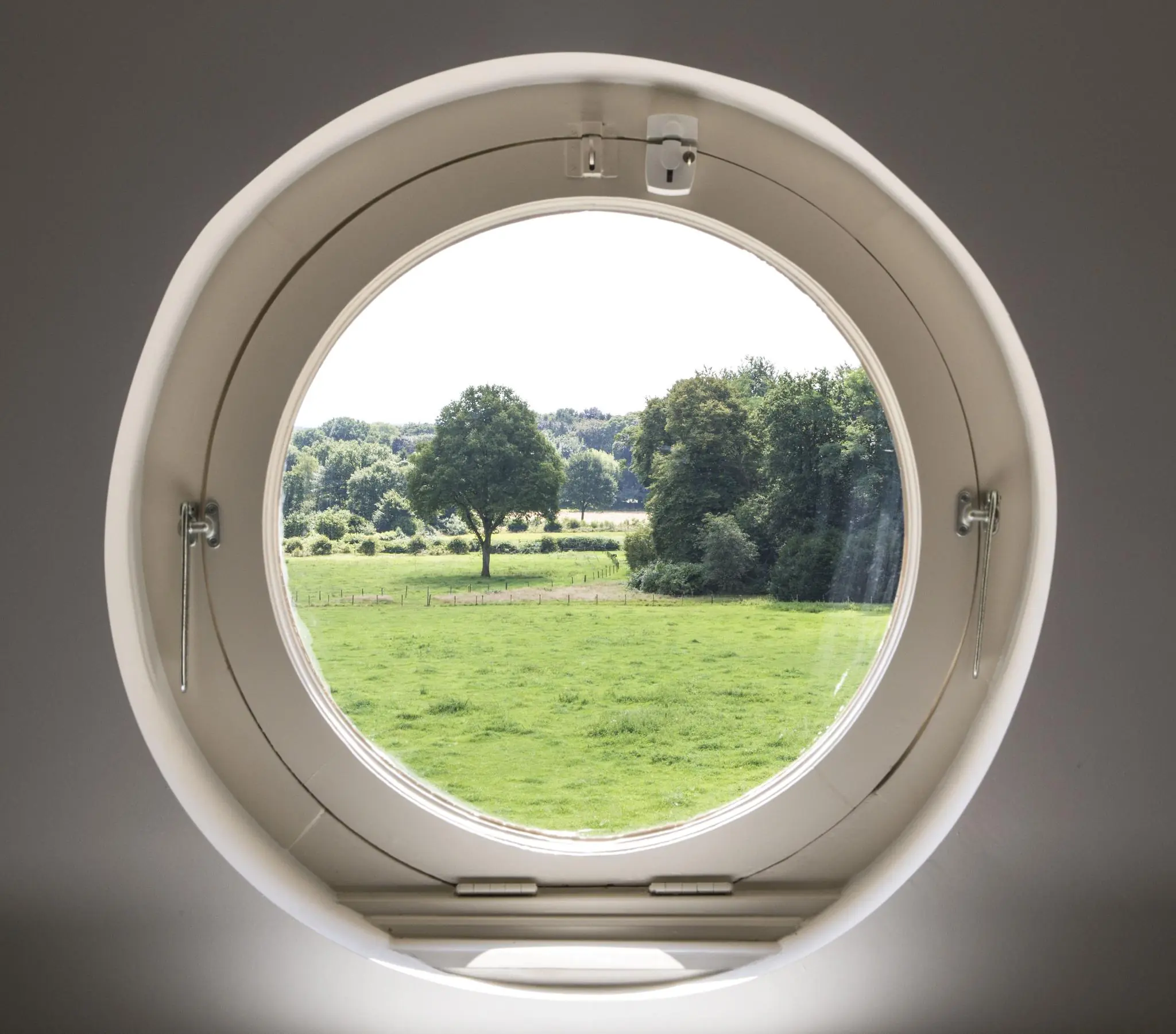 Uitzicht op een groen landschap met bomen, gezien door een rond raam aan de Naarderstraat.