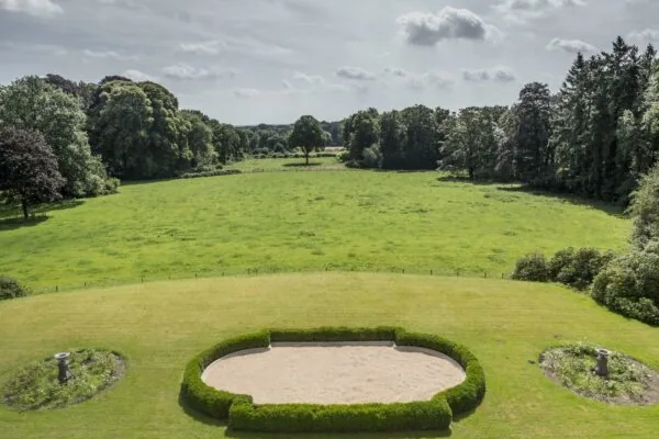 Uitzicht op een weids groen veld met een symmetrisch aangelegde tuin aan de Naarderstraat, omringd door bomen.