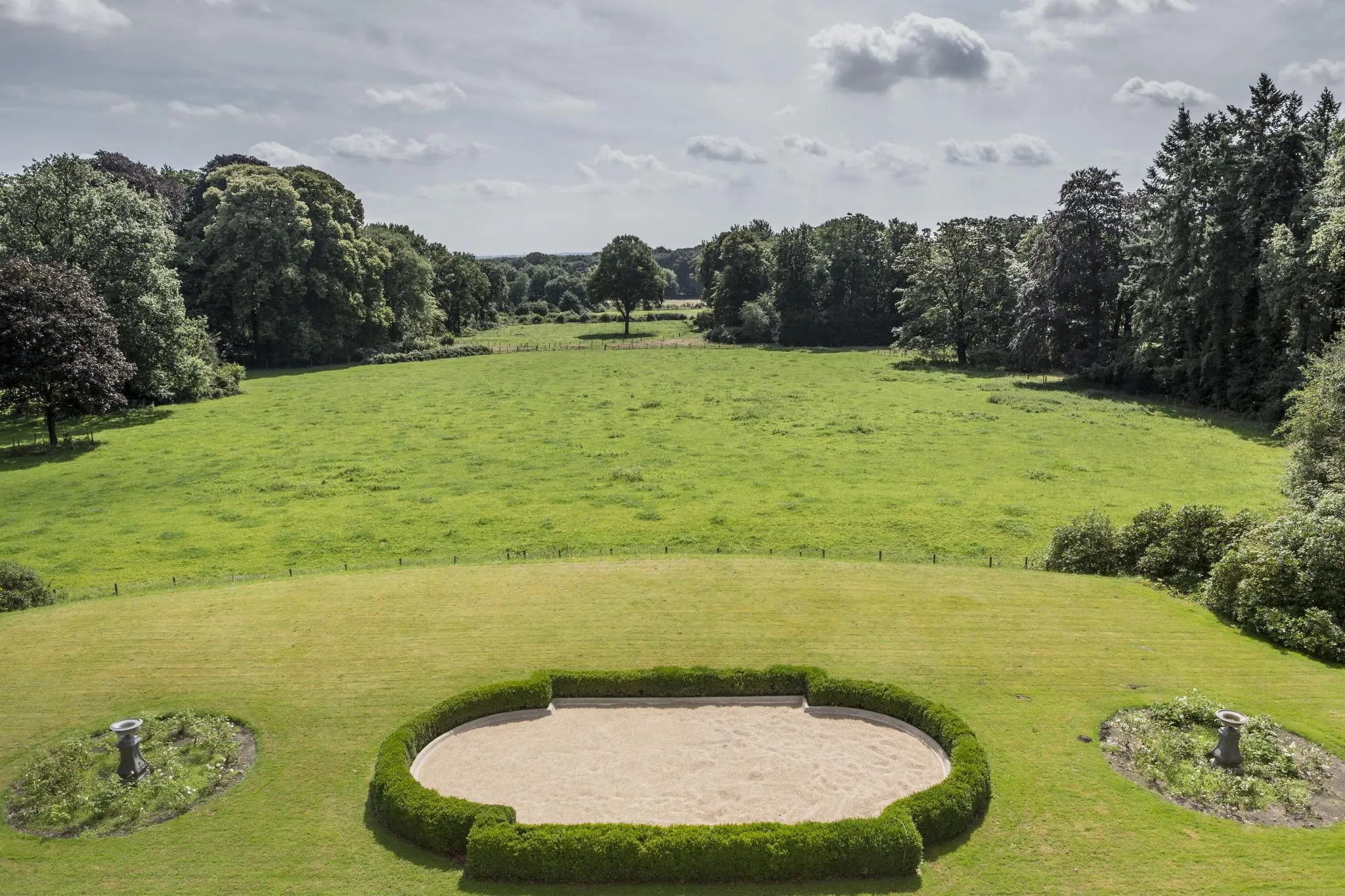 Uitzicht op een weids groen veld met een symmetrisch aangelegde tuin aan de Naarderstraat, omringd door bomen.