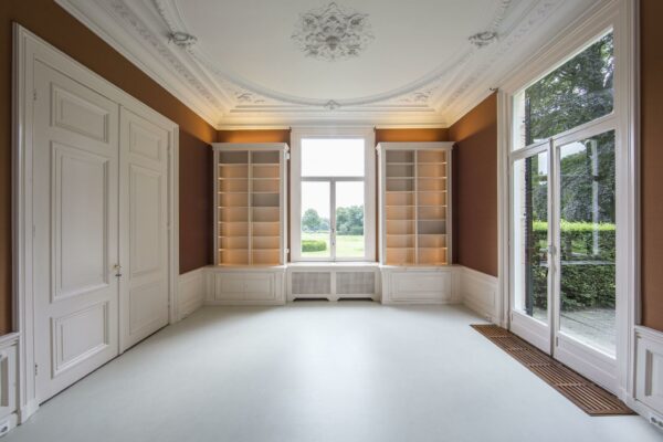 Klassieke, lege kamer aan de Naarderstraat met wit plafondornament, ingebouwde boekenkasten en uitzicht op een groene tuin.