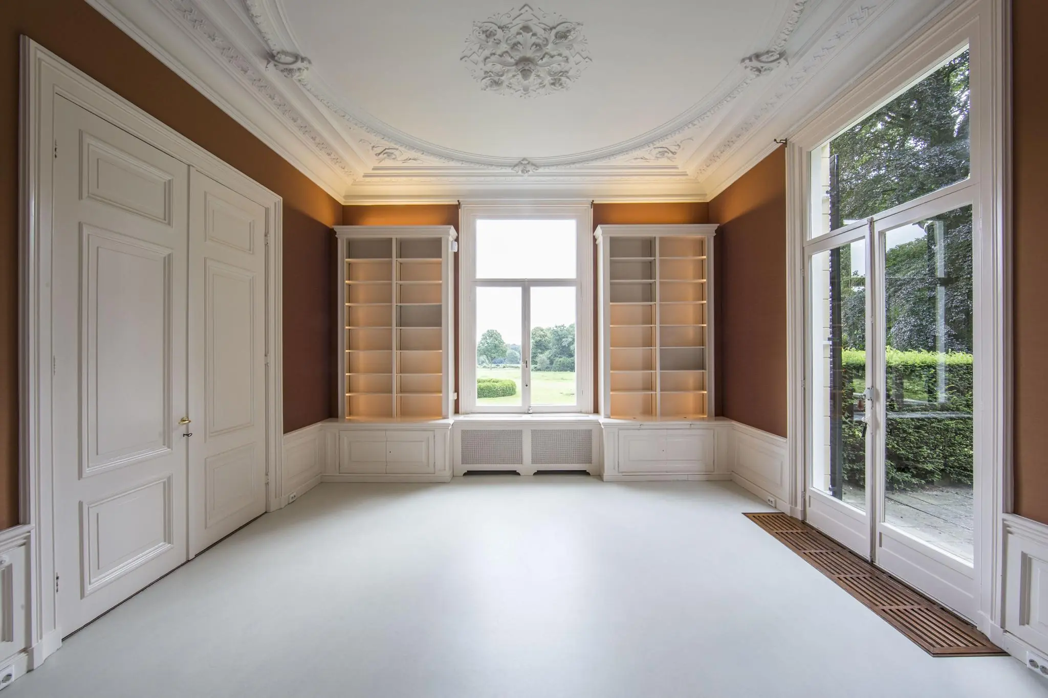 Klassieke, lege kamer aan de Naarderstraat met wit plafondornament, ingebouwde boekenkasten en uitzicht op een groene tuin.