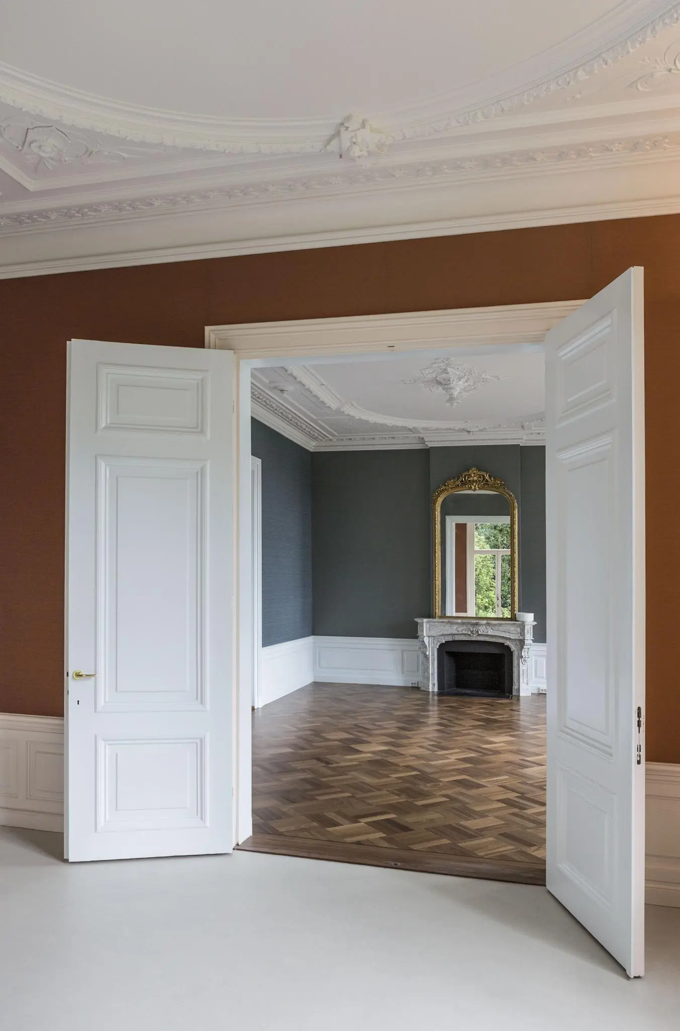 Doorkijk naar een stijlvolle kamer aan de Naarderstraat met visgraatparket, sierplafond, marmeren schouw en grote spiegel.