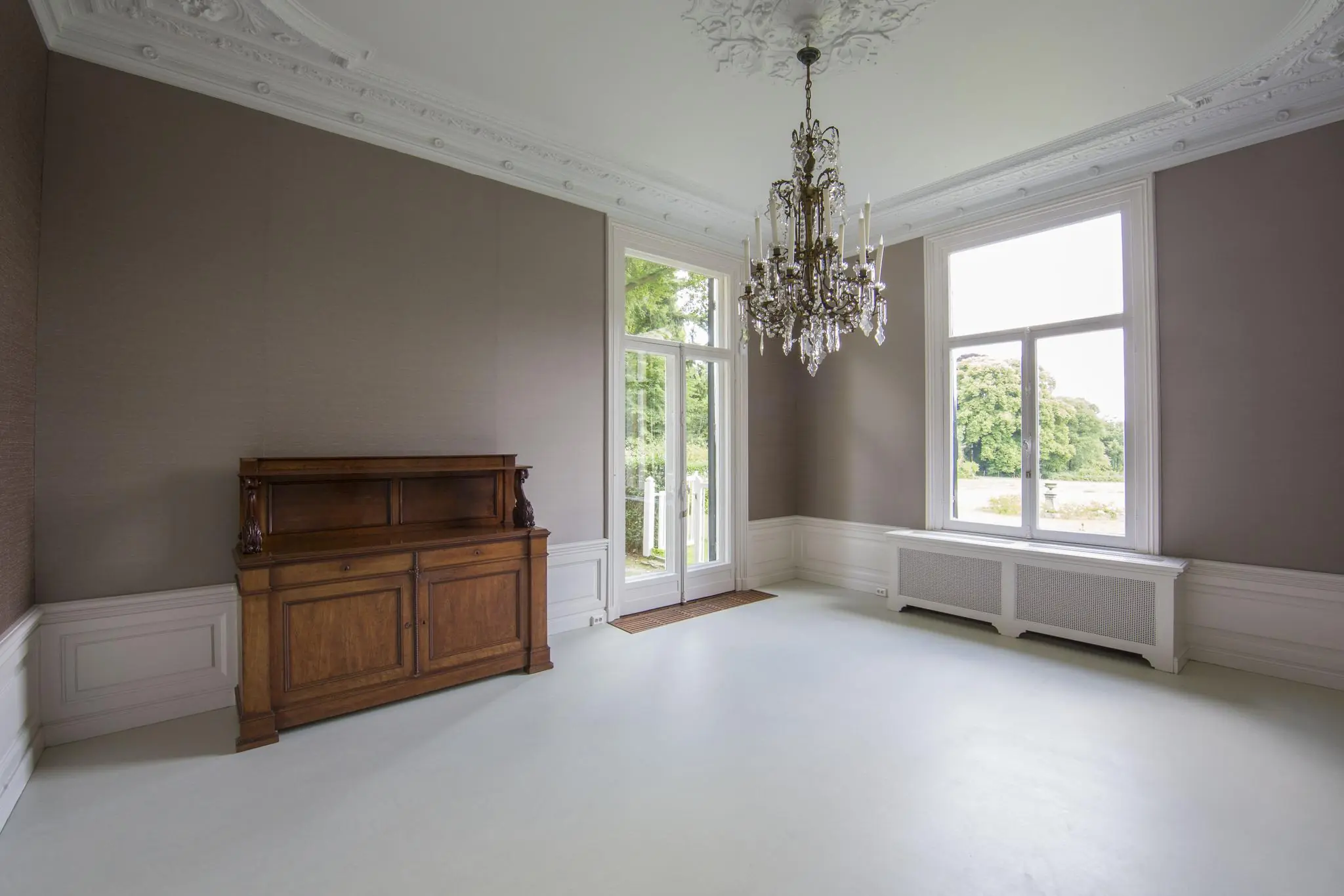Lichte, klassieke kamer aan de Naarderstraat met hoog plafond, ornamenten, kroonluchter en houten dressoir.