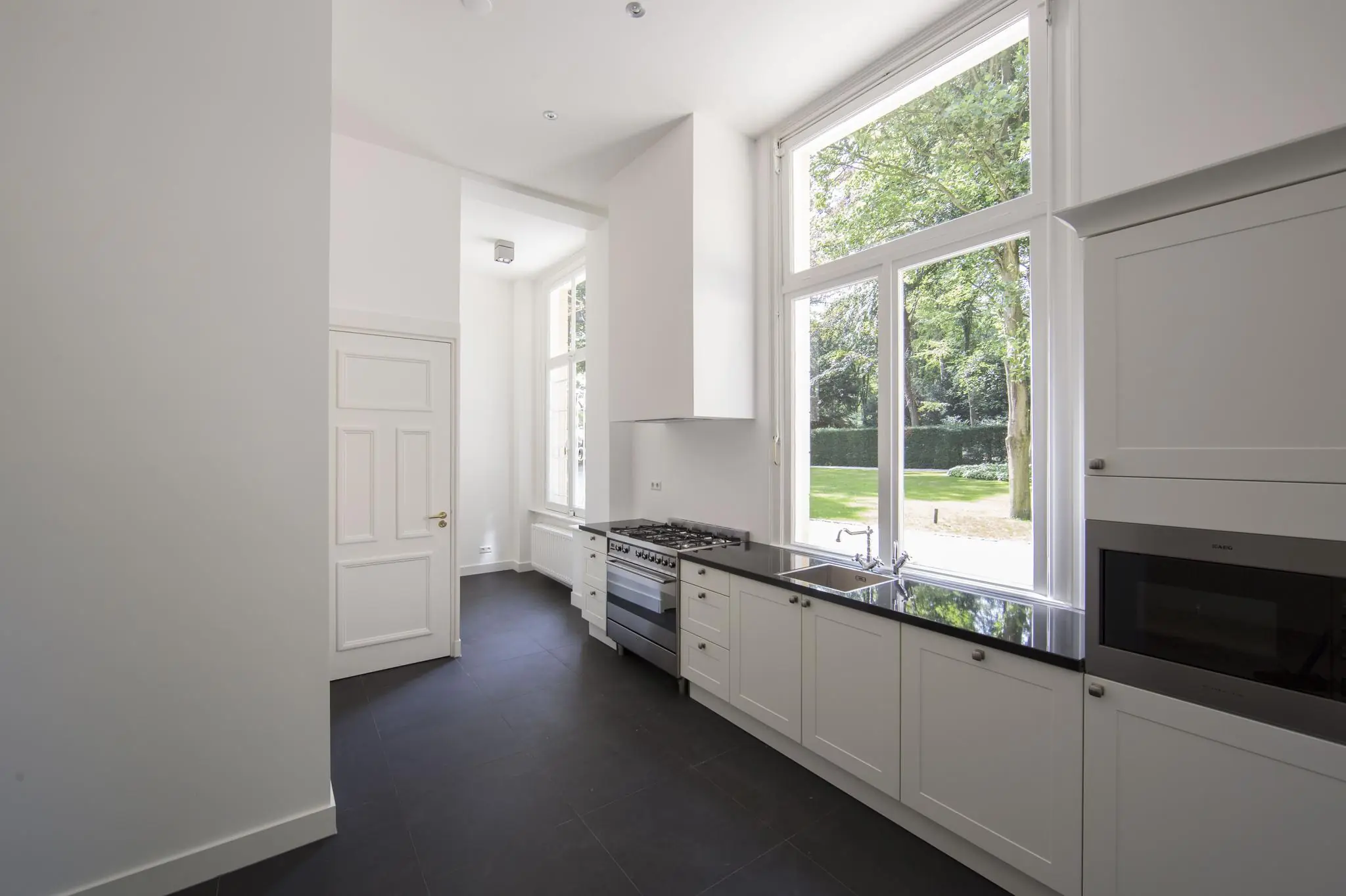 Moderne keuken aan de Naarderstraat met wit interieur, zwarte vloer en groot raam met uitzicht op groene tuin.
