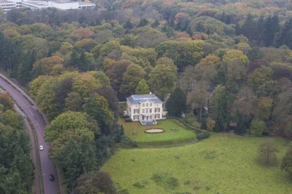 Luchtfoto van een grote, witte villa aan de Naarderstraat omringd door bosrijk gebied en een ruime tuin.