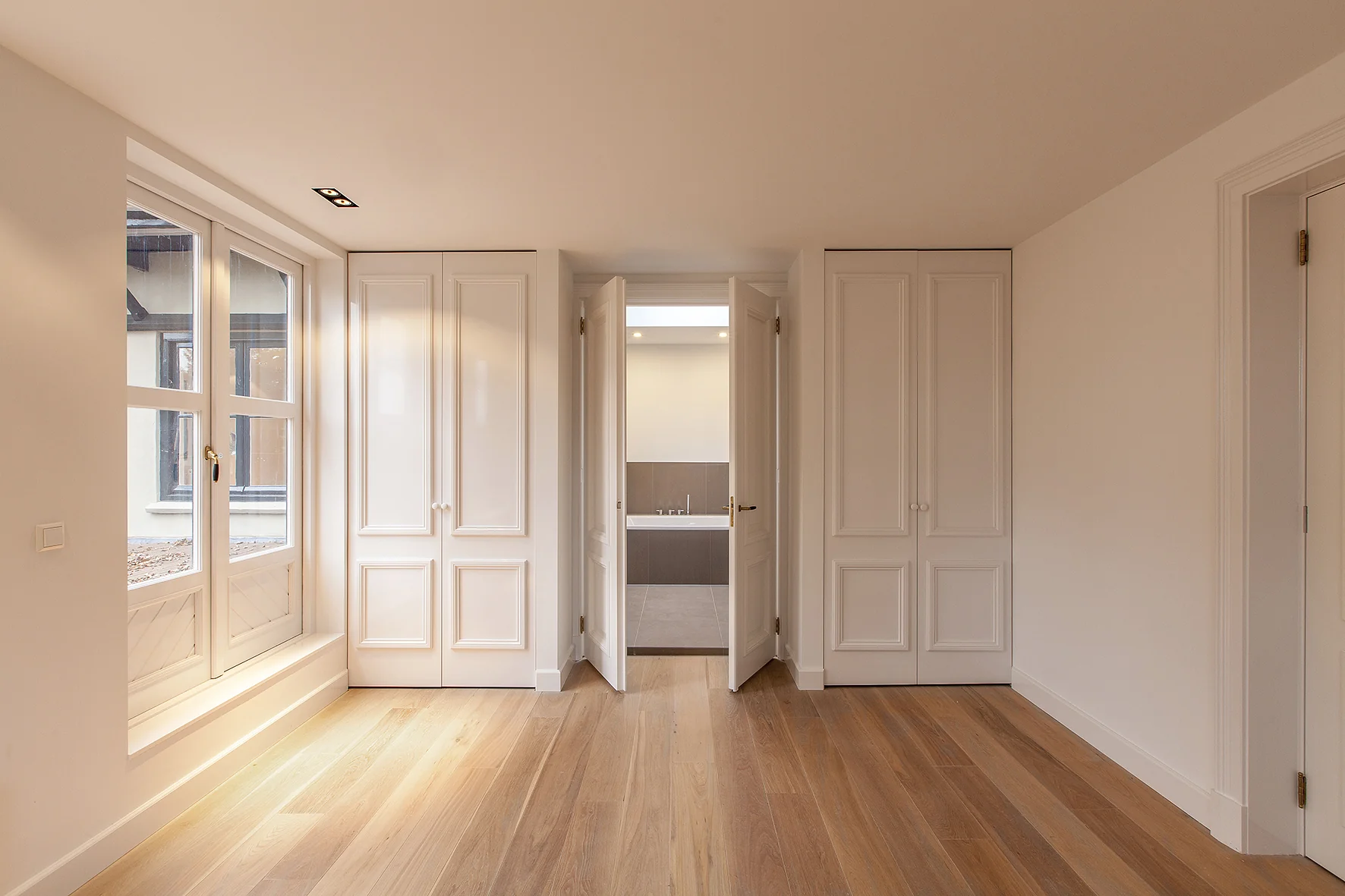 Lichte slaapkamer met houten vloer, inbouwkasten en dubbele deuren naar een moderne badkamer aan de Naarderstraat.